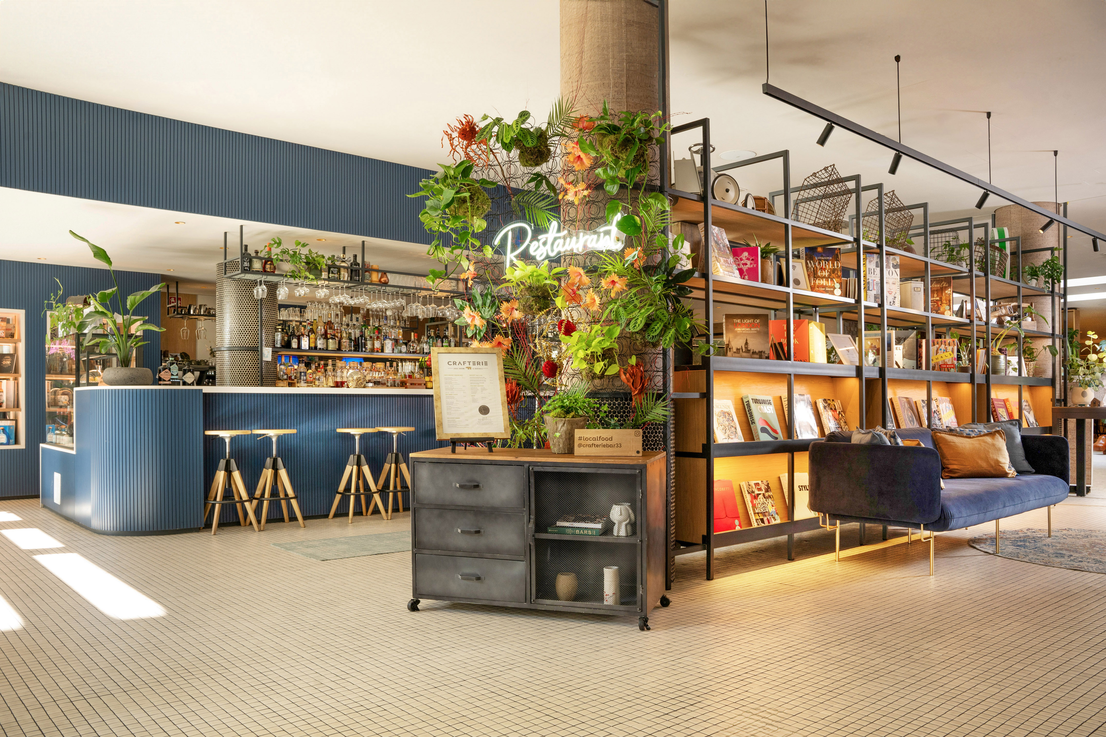 Blue bar and restaurant lounge with neon sign, lush plants, and bookshelves.
