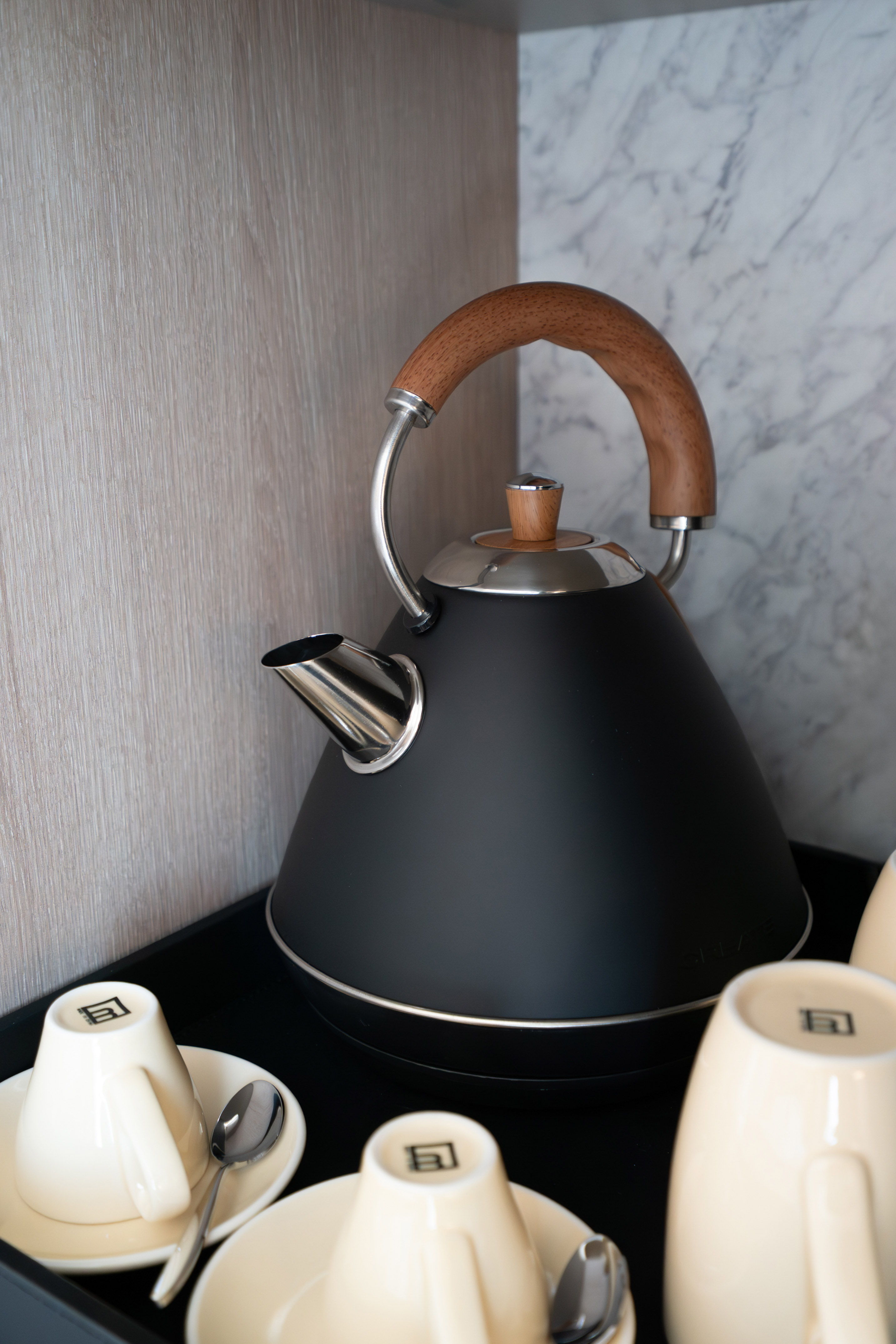Black modern kettle with wood handle, cream cups, and spoons on a black tray.