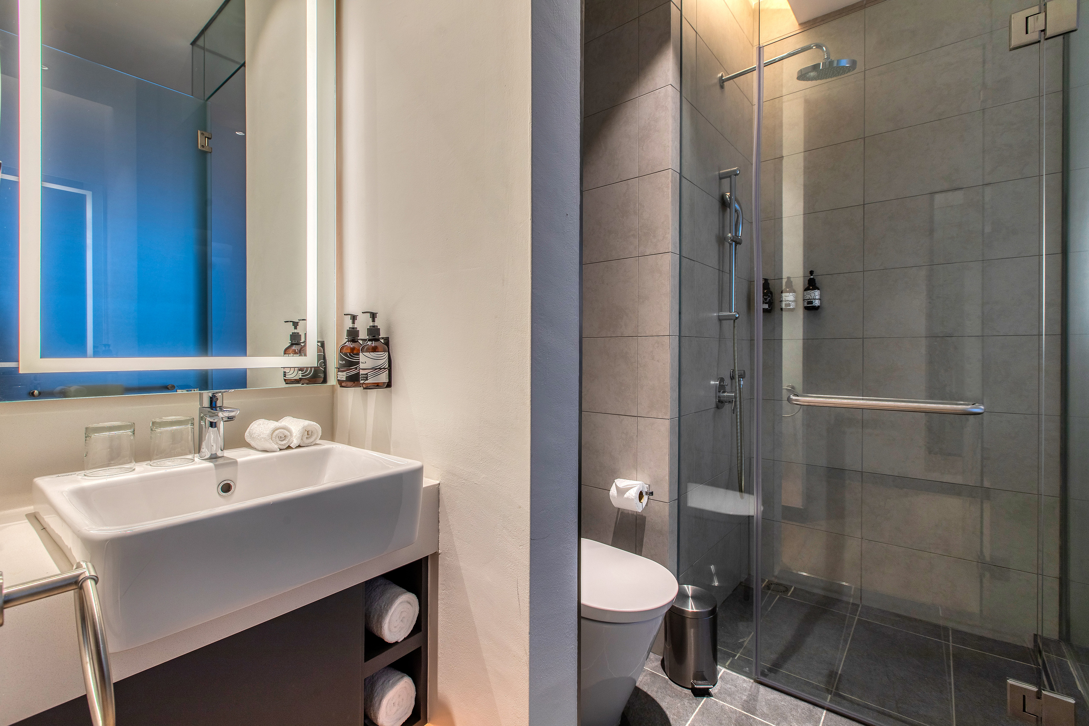 Modern hotel bathroom with illuminated mirror, sink, toilet, and tiled walk-in shower.