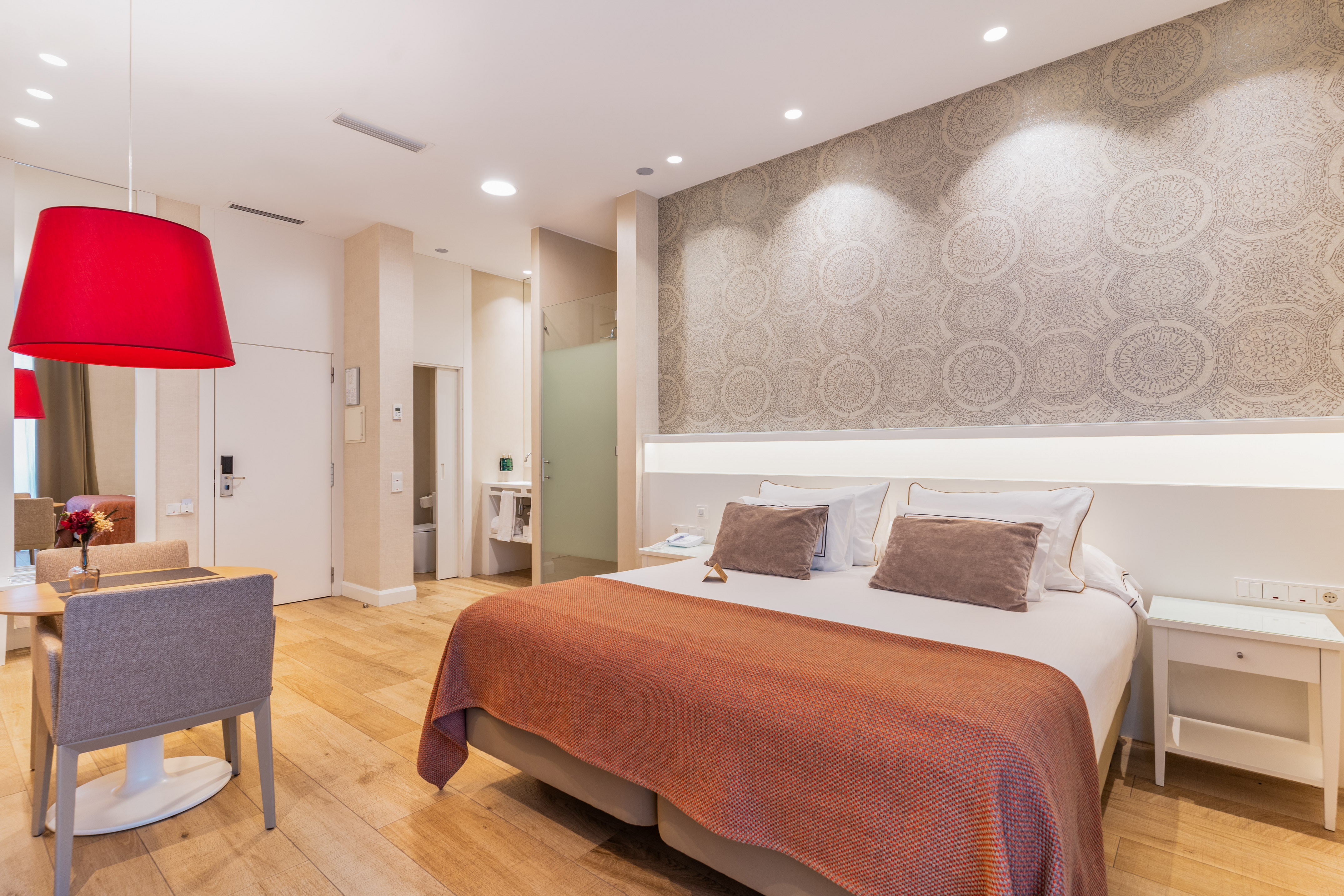 Modern hotel suite with king bed, patterned wall, red lamp, and wooden floor.