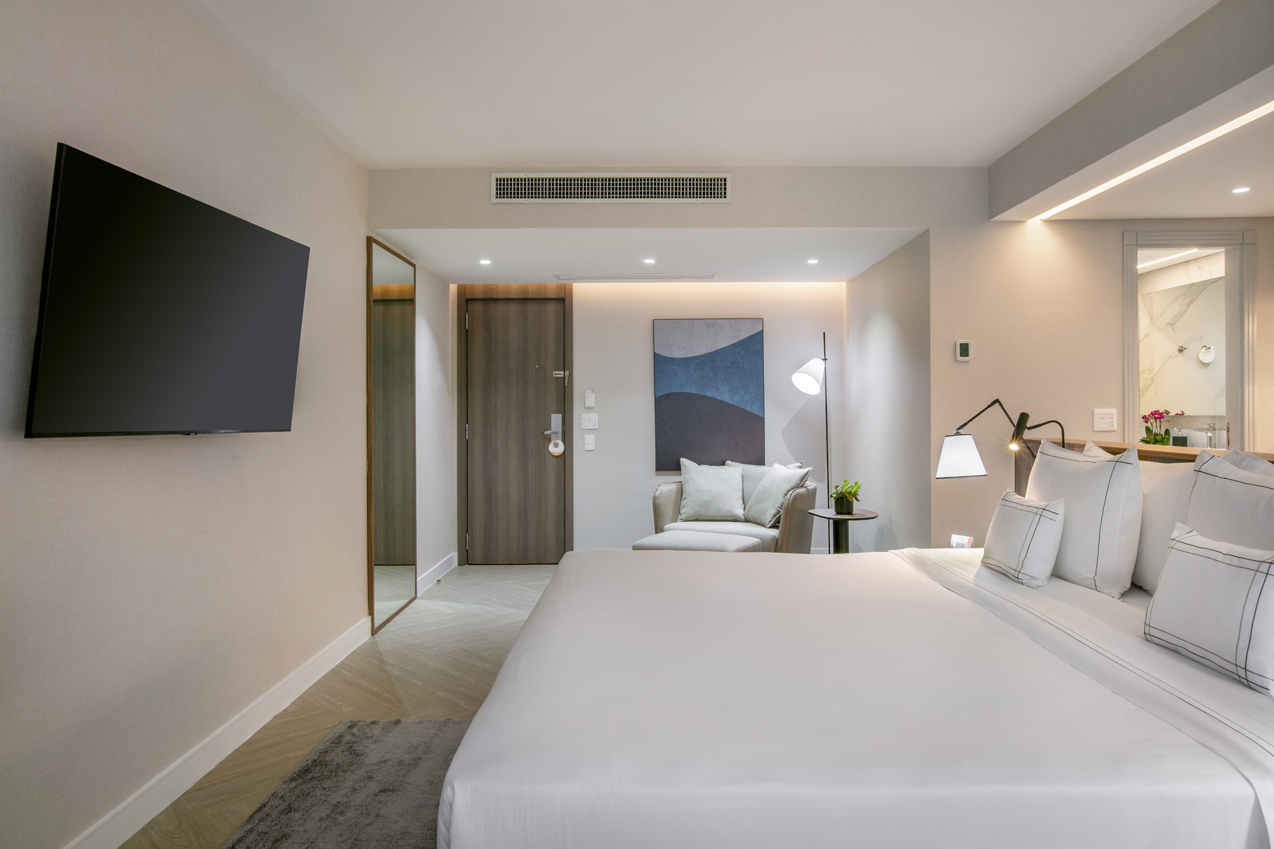 Modern hotel bedroom with large white bed, wall-mounted TV, and chic lounge area.