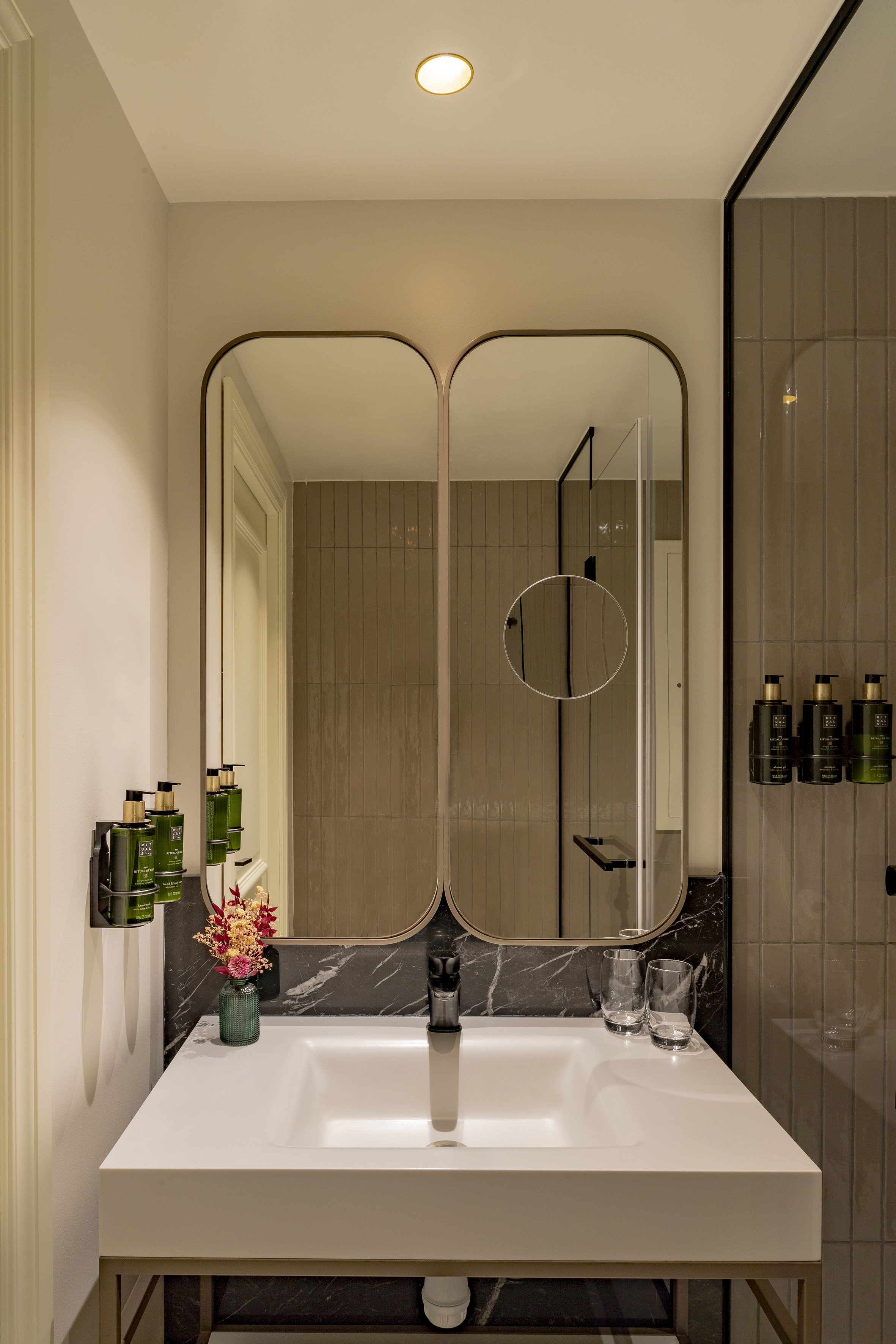 Modern bathroom vanity: white sink, black marble, arched mirrors, shower view, hotel toiletries.