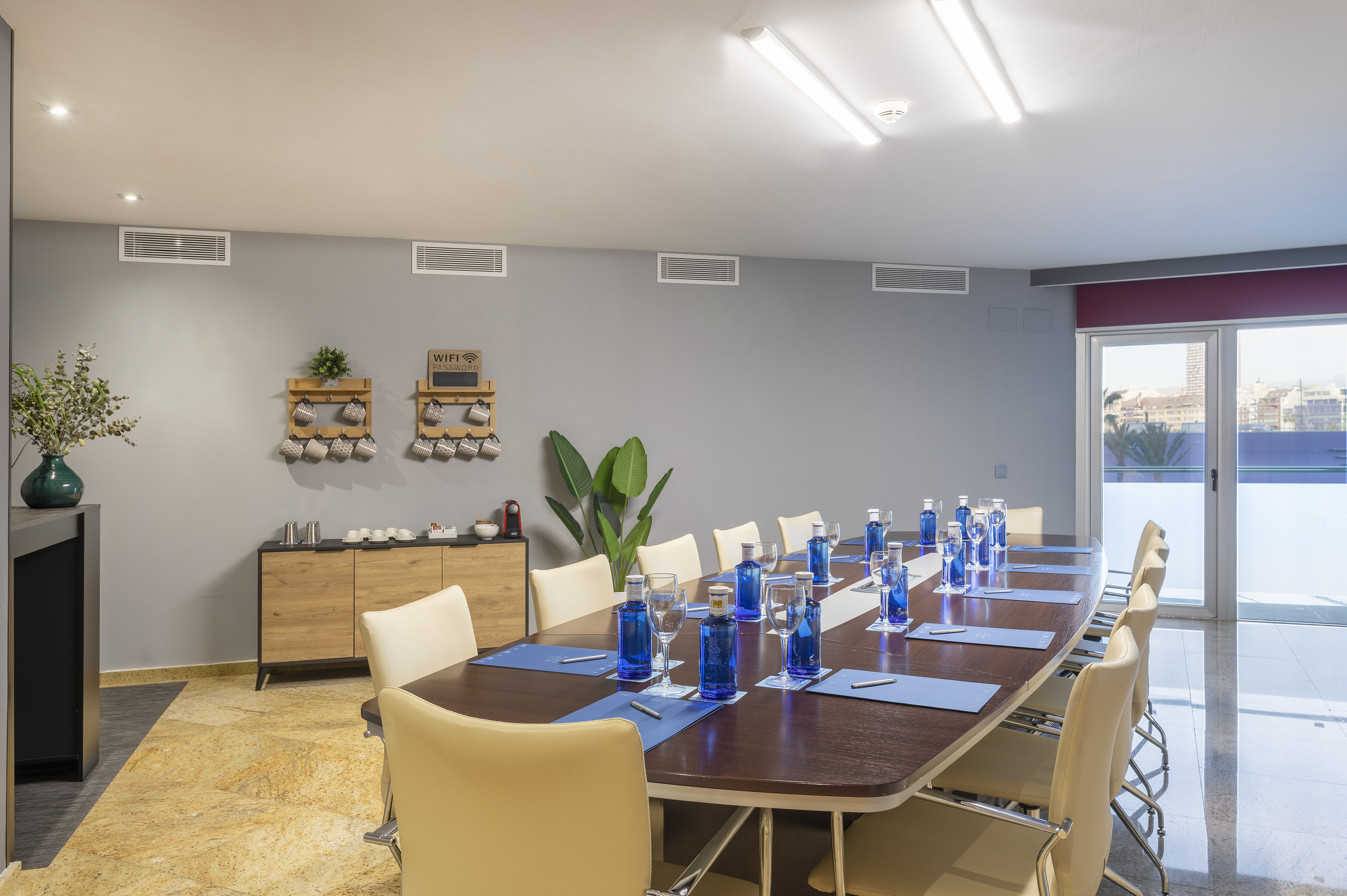 Modern conference room with large table, beige chairs, refreshments, city views.