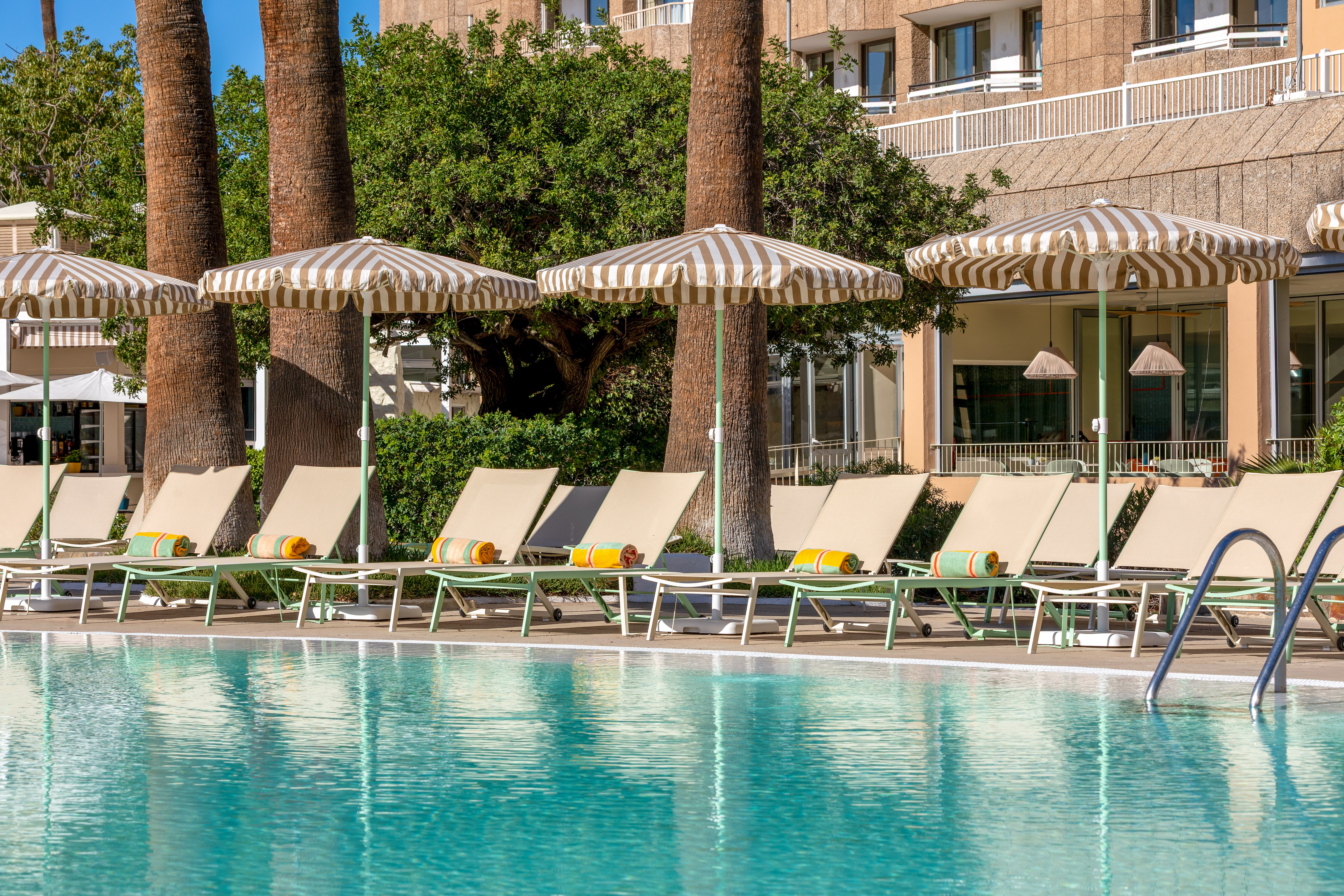 Resort swimming pool with lounge chairs, striped umbrellas, and palm trees.