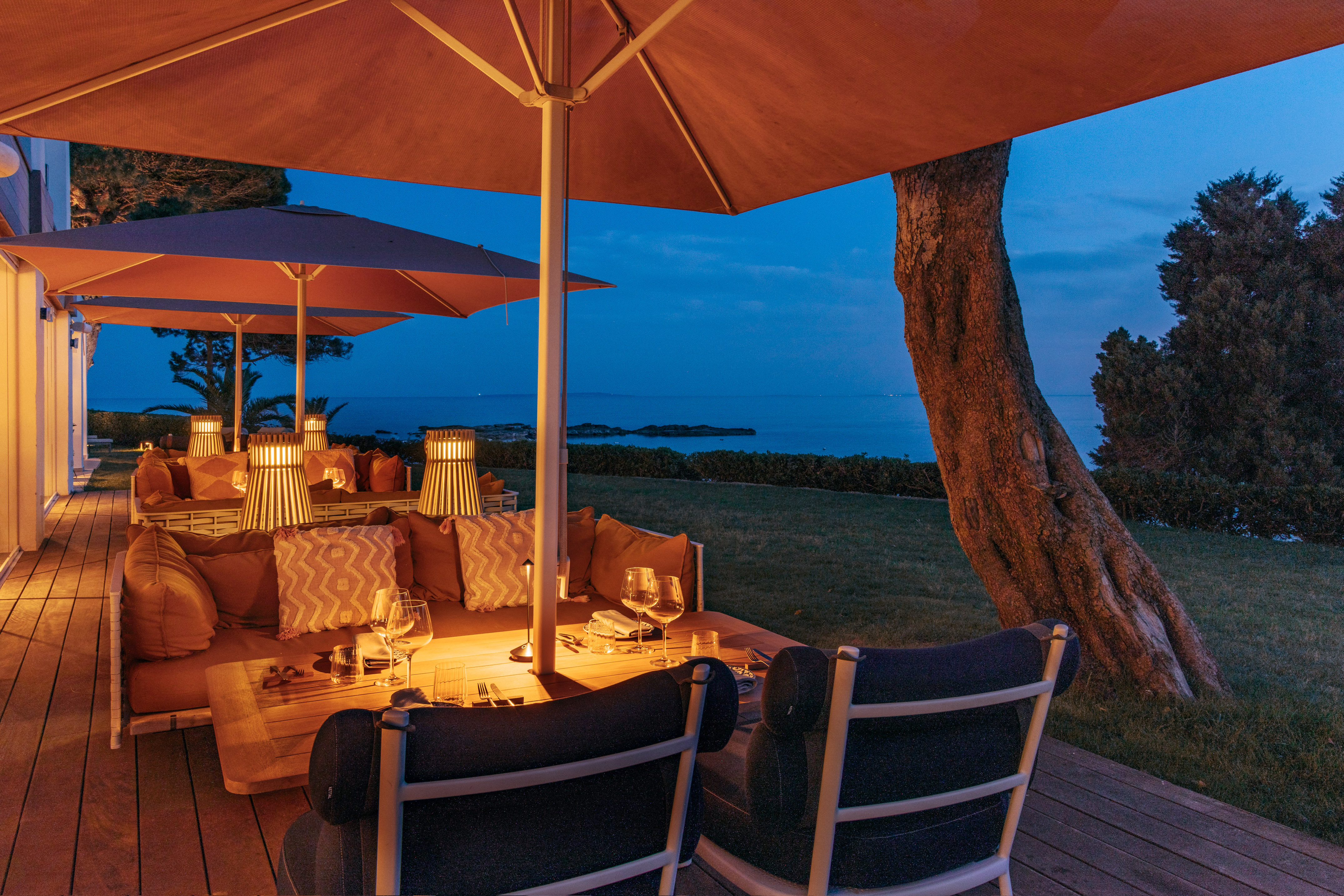 Outdoor restaurant patio at dusk, ocean view, cushioned seating, and table lamps.