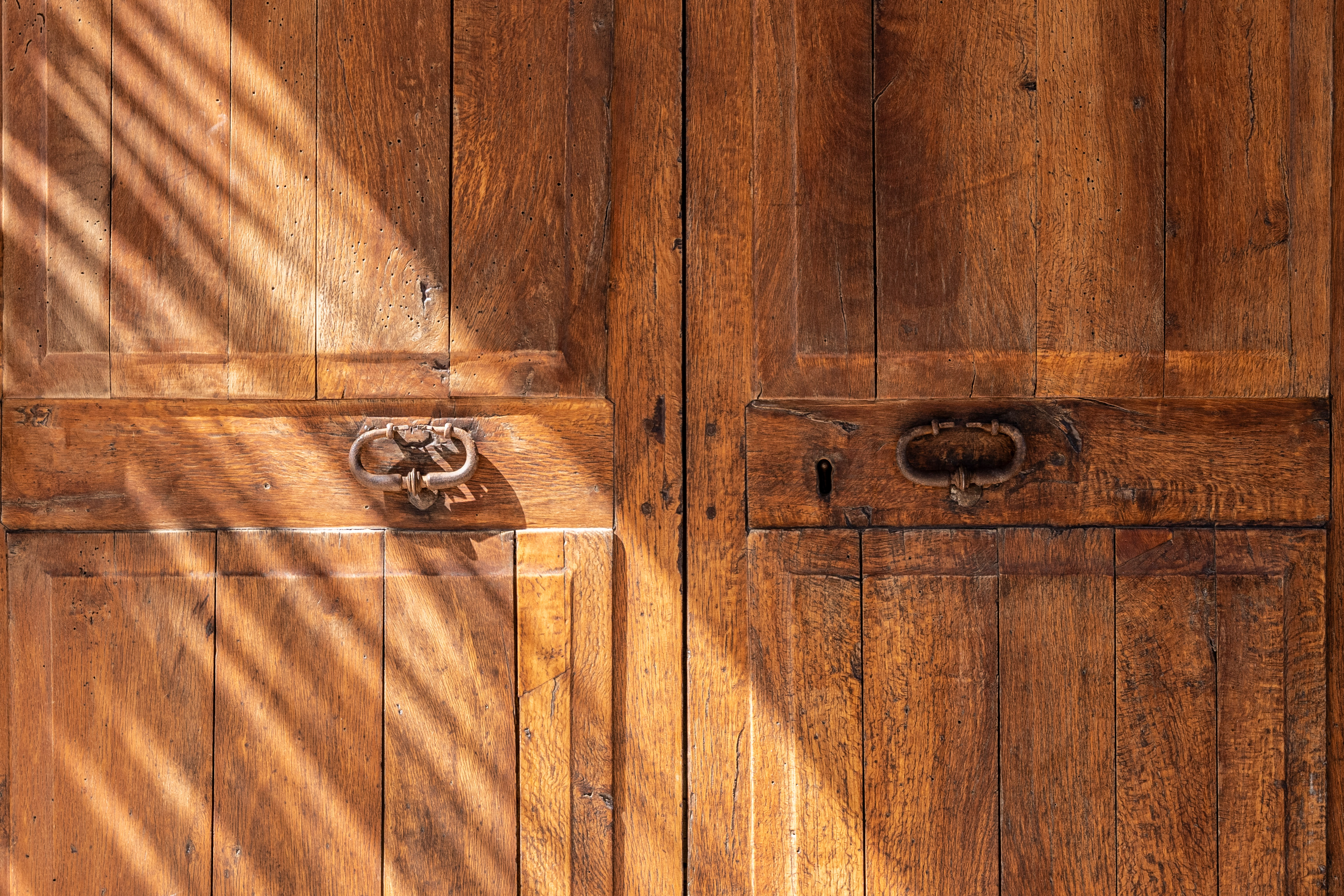 Rustic wooden double door with antique metal knockers, keyhole, and sunlight stripes.