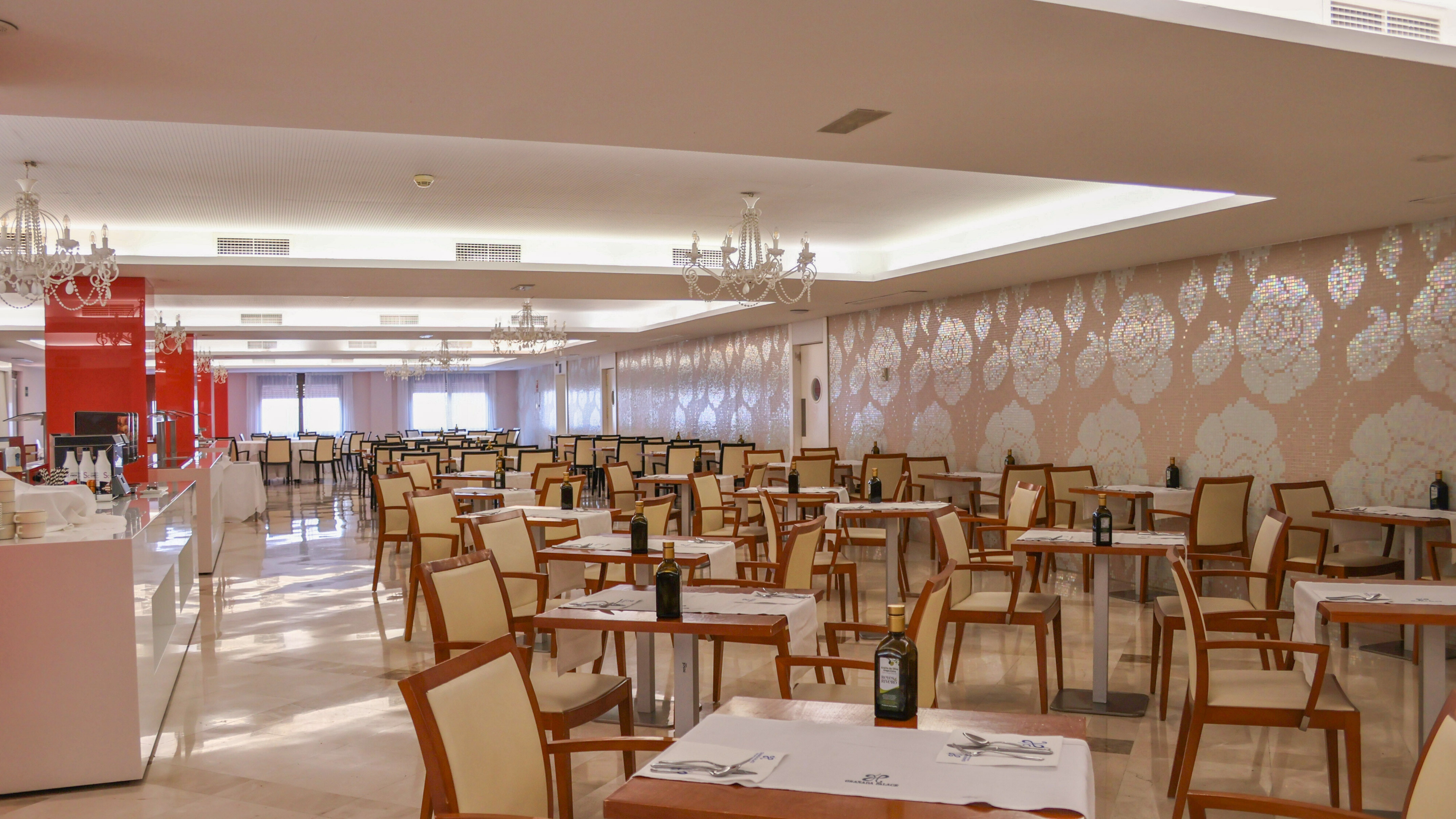 Spacious hotel restaurant with set tables, chandeliers, and floral mosaic wall.