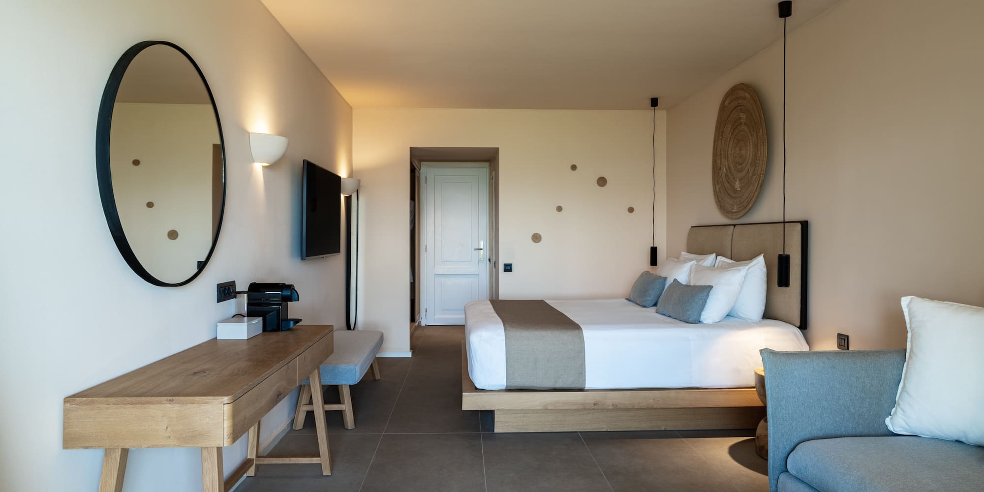 Modern hotel suite with neutral decor, wooden desk, large bed, and grey sofa.