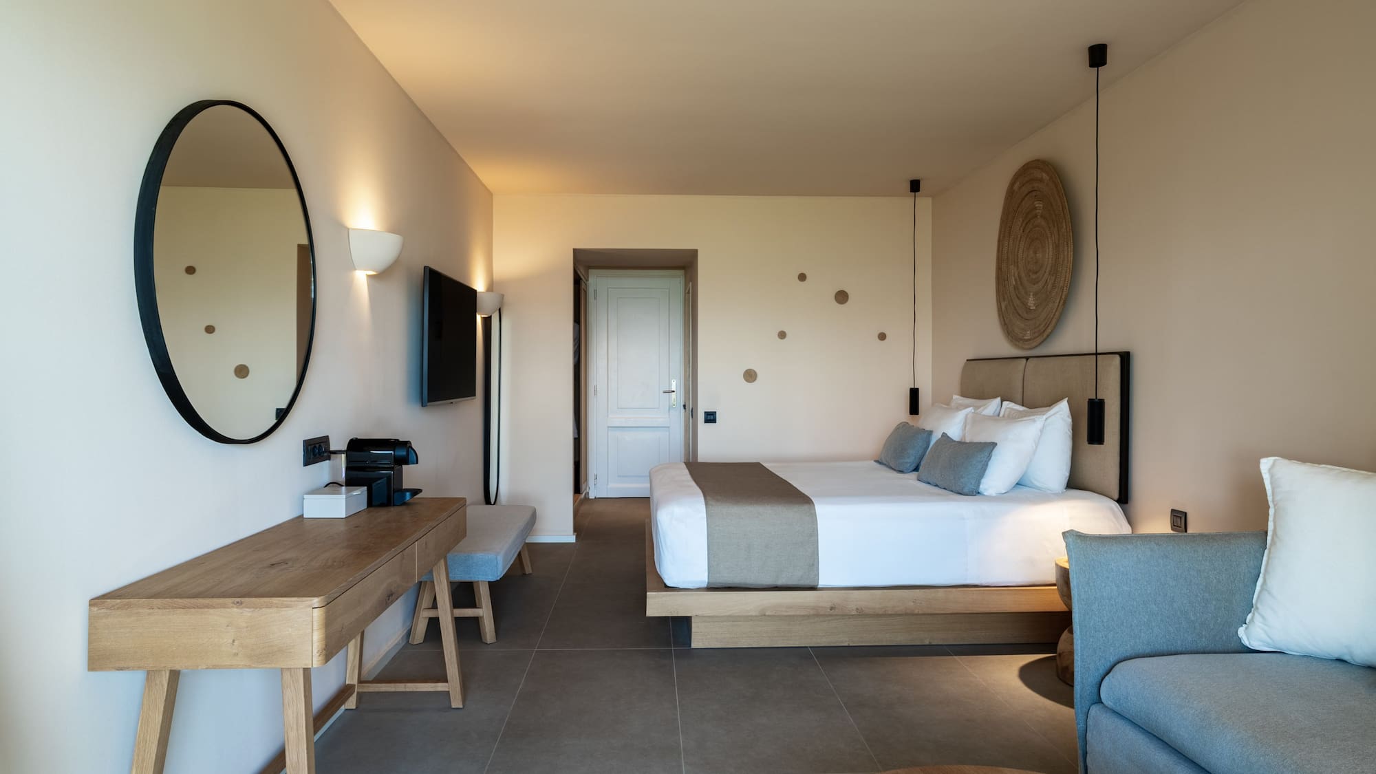 Modern hotel suite with neutral decor, wooden desk, large bed, and grey sofa.