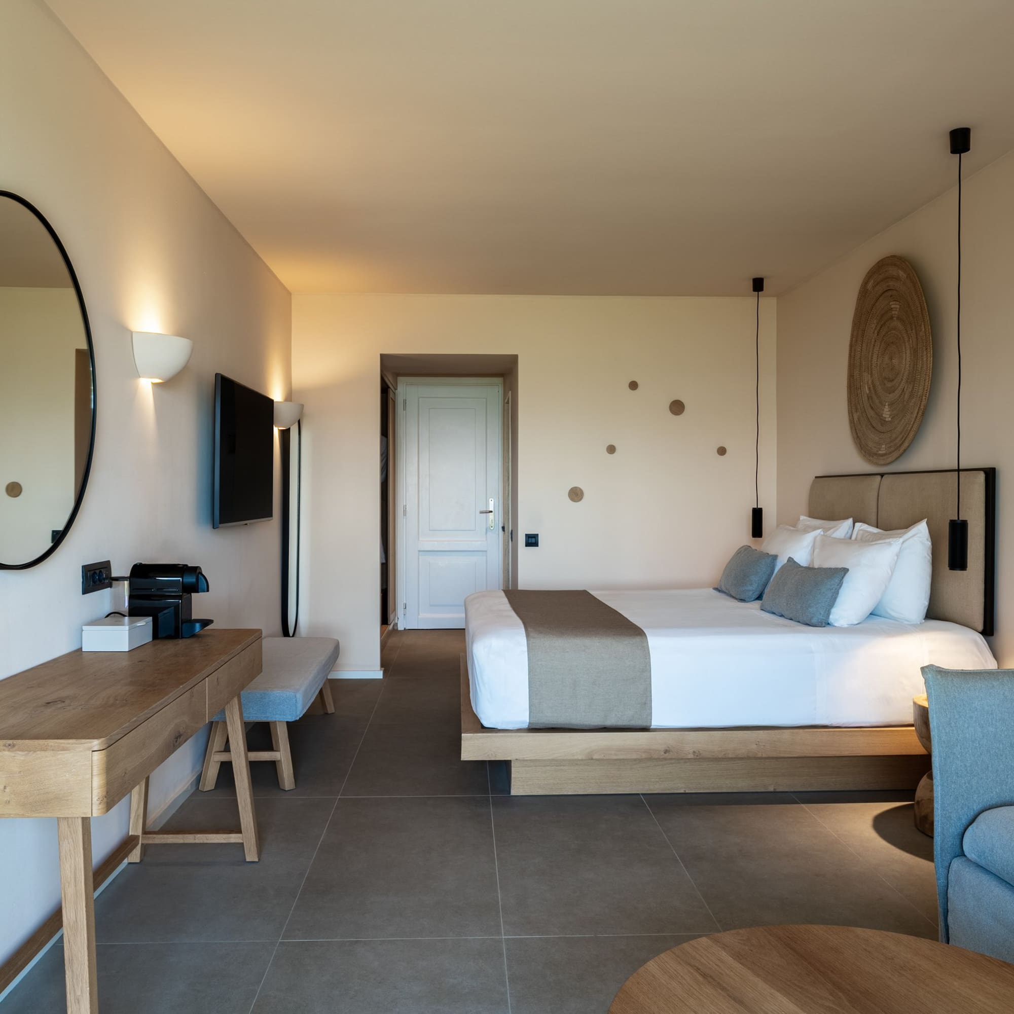 Modern hotel suite with neutral decor, wooden desk, large bed, and grey sofa.