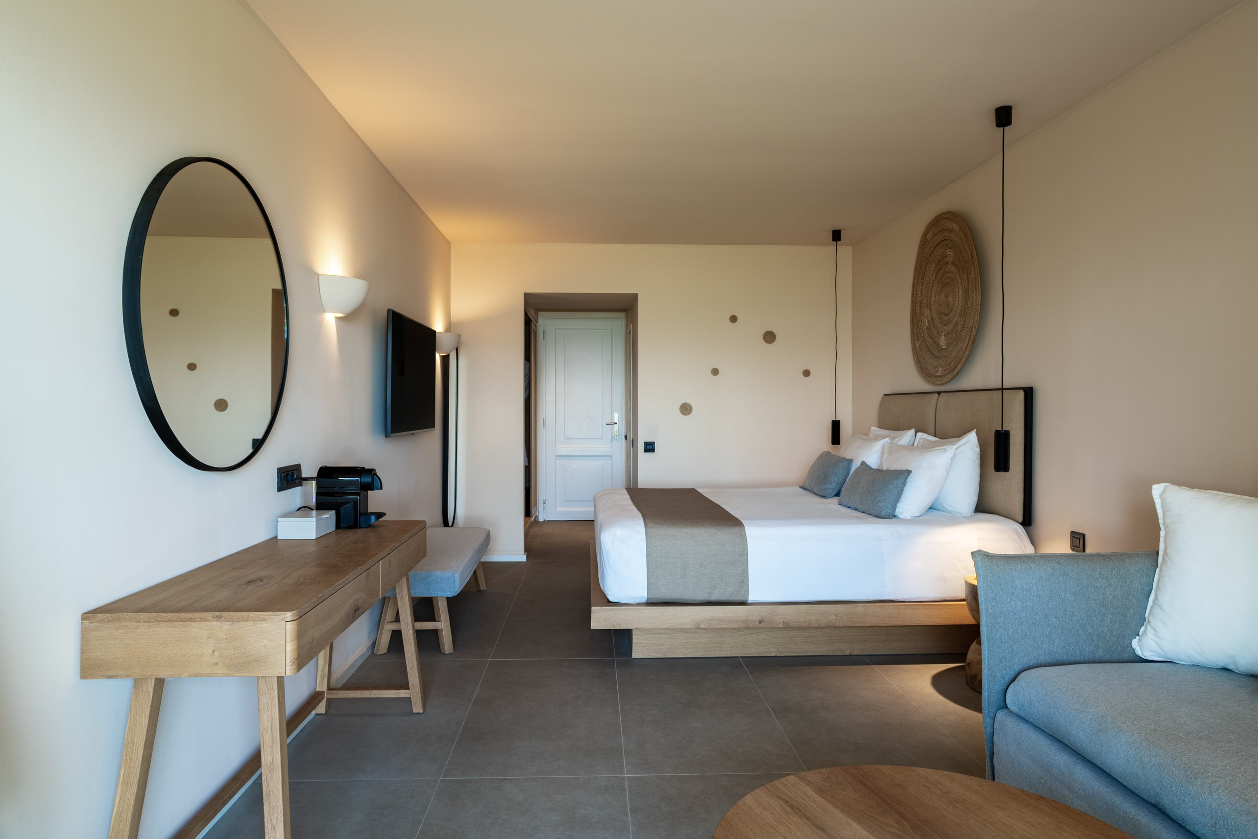 Modern hotel suite with neutral decor, wooden desk, large bed, and grey sofa.