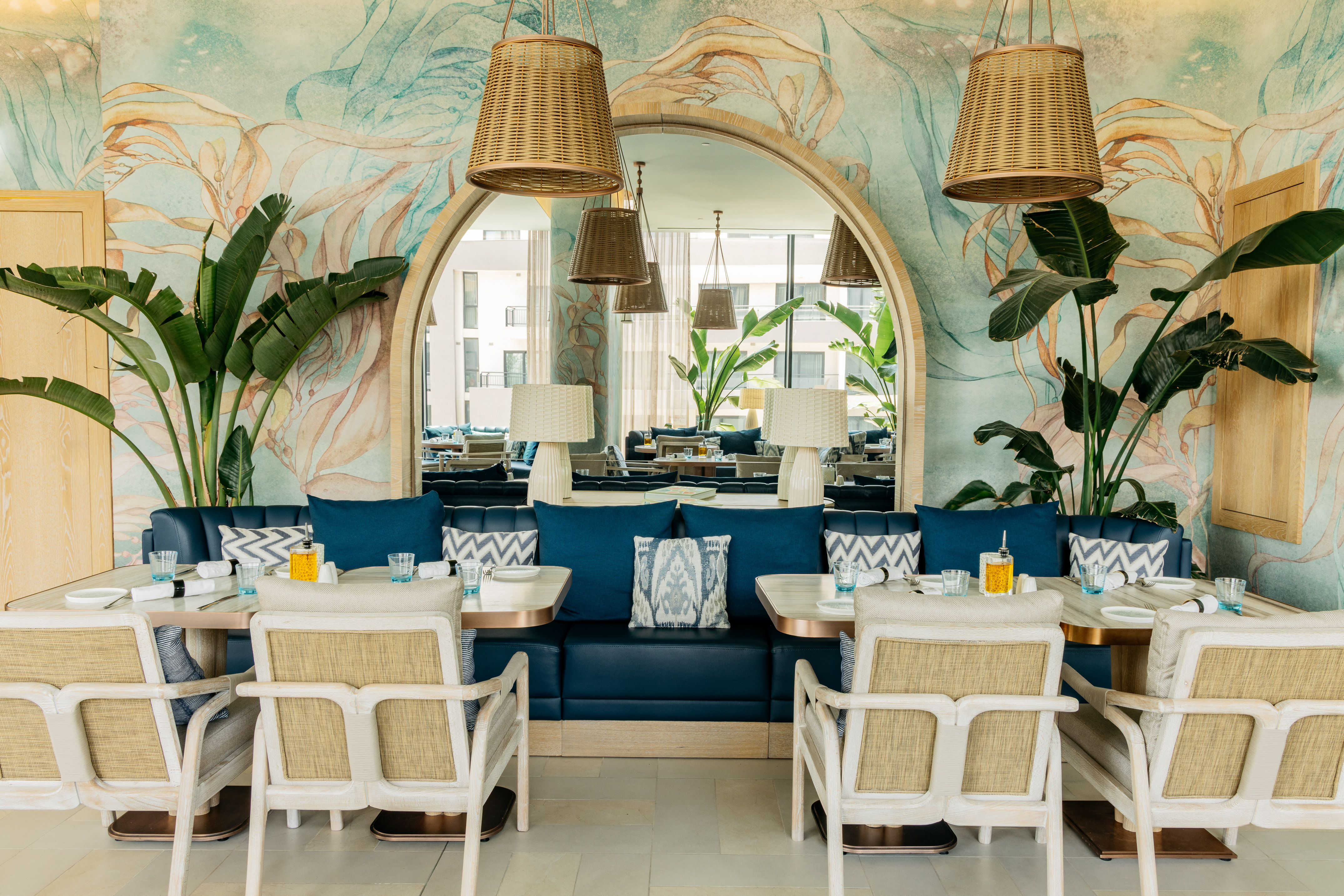 Coastal restaurant with blue banquette, tropical wallpaper, and rattan lights.