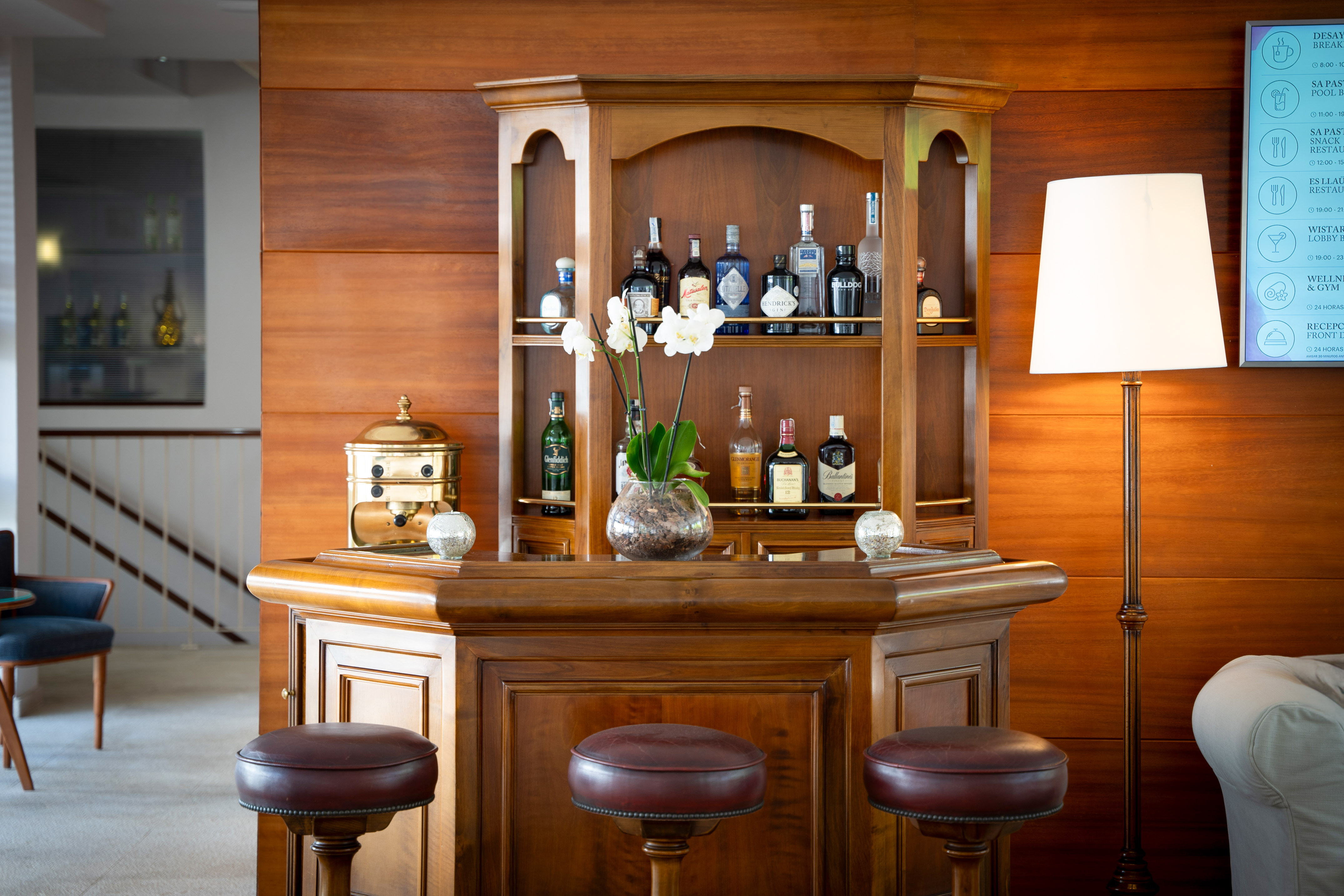 Wooden bar with spirits, three leather stools, and orchids in a hotel lounge.
