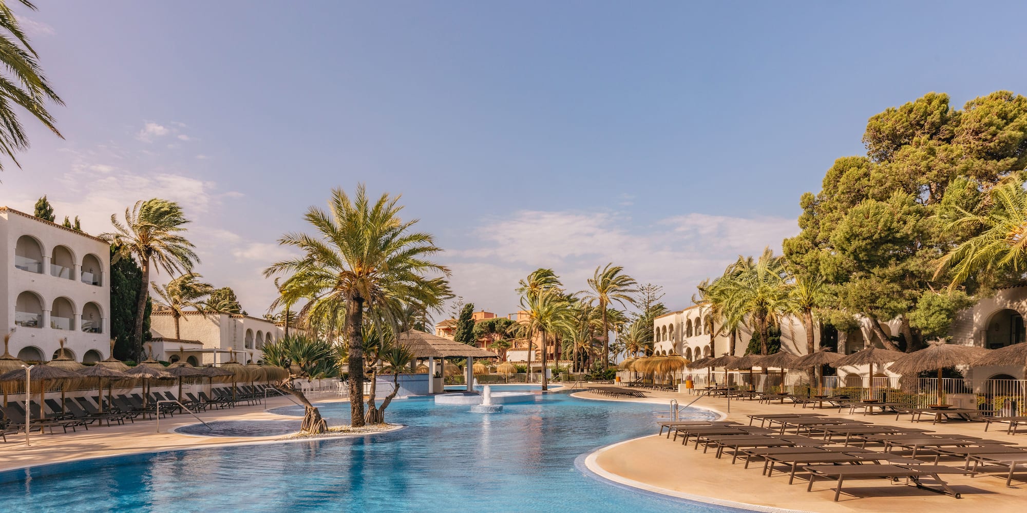 Tropical resort pool, palm trees, sunbeds, and hotel buildings under a clear sky.