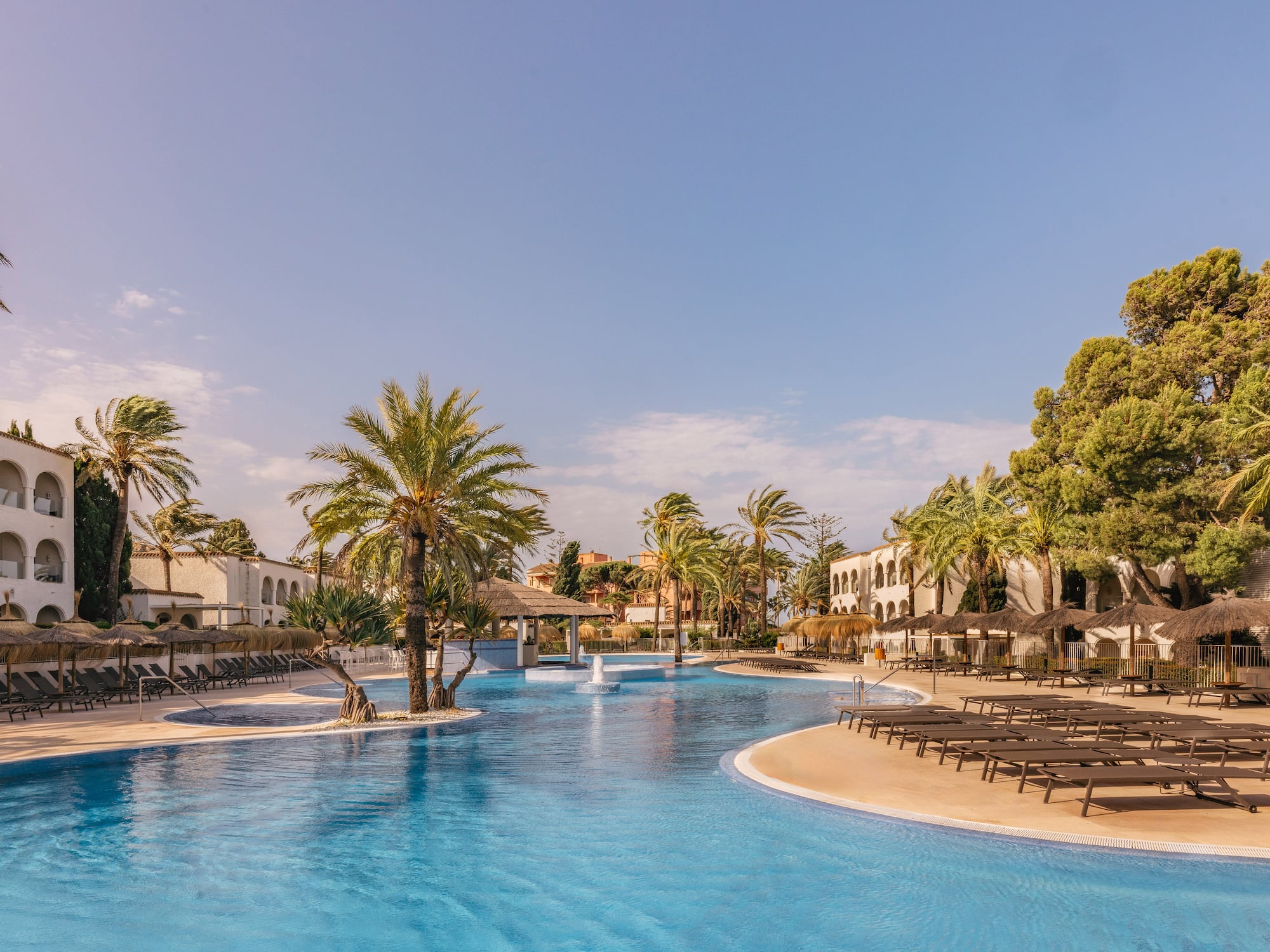 Tropical resort pool, palm trees, sunbeds, and hotel buildings under a clear sky.