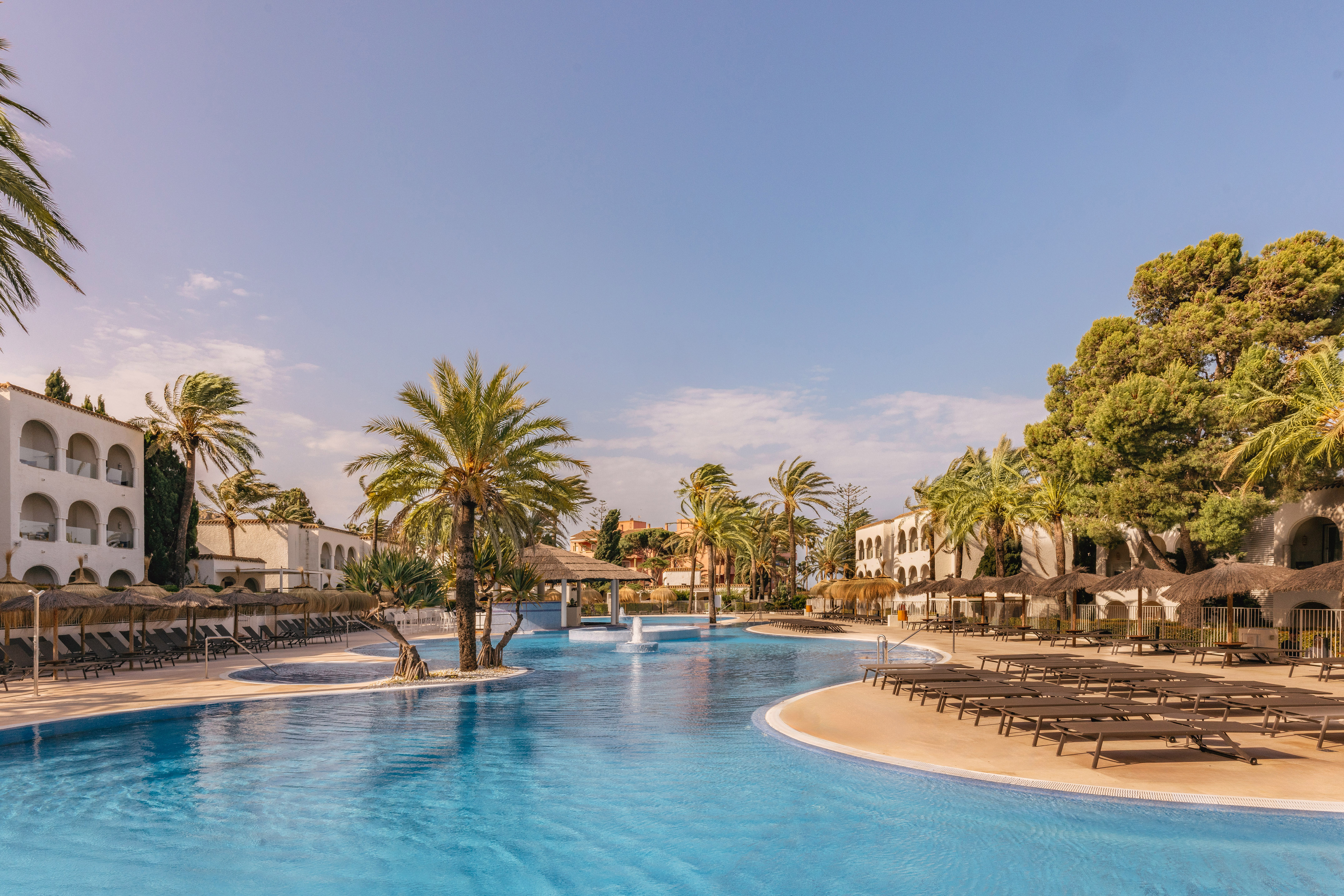 Blue tropical resort pool with palm trees, sun loungers, and hotel buildings.