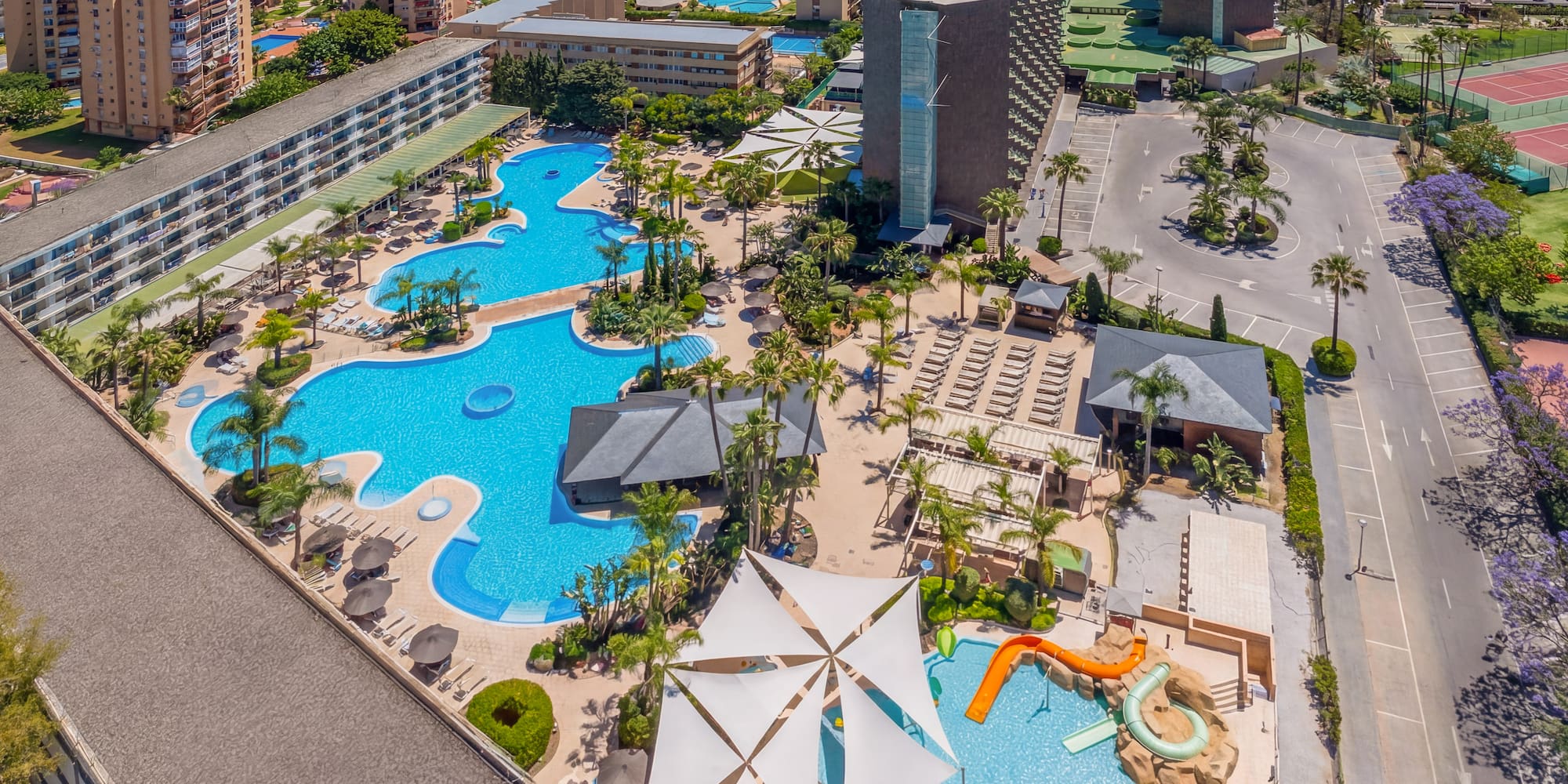 Aerial resort view: hotel, multiple pools, water slides, palm trees, beach, ocean.