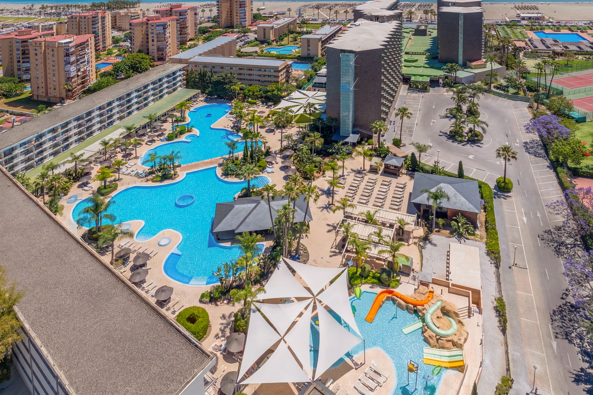 Aerial resort view: hotel, multiple pools, water slides, palm trees, beach, ocean.