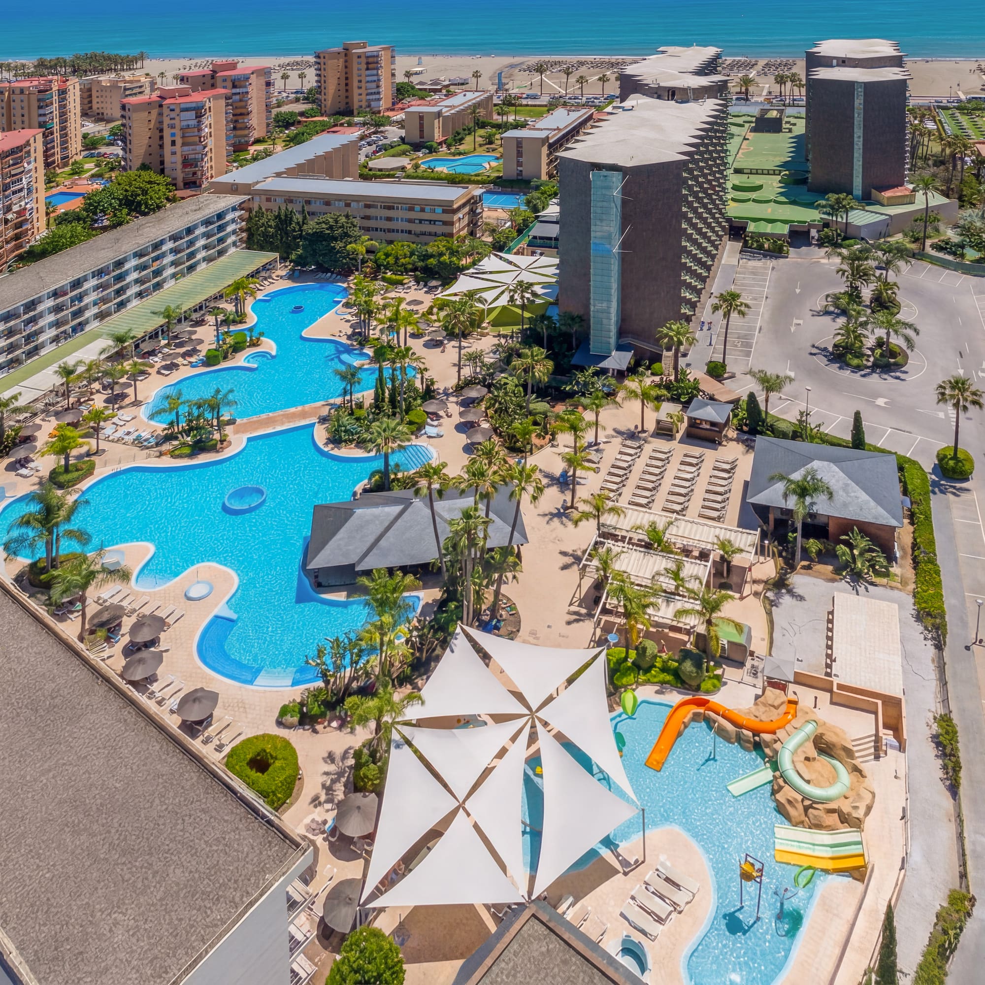 Aerial resort view: hotel, multiple pools, water slides, palm trees, beach, ocean.
