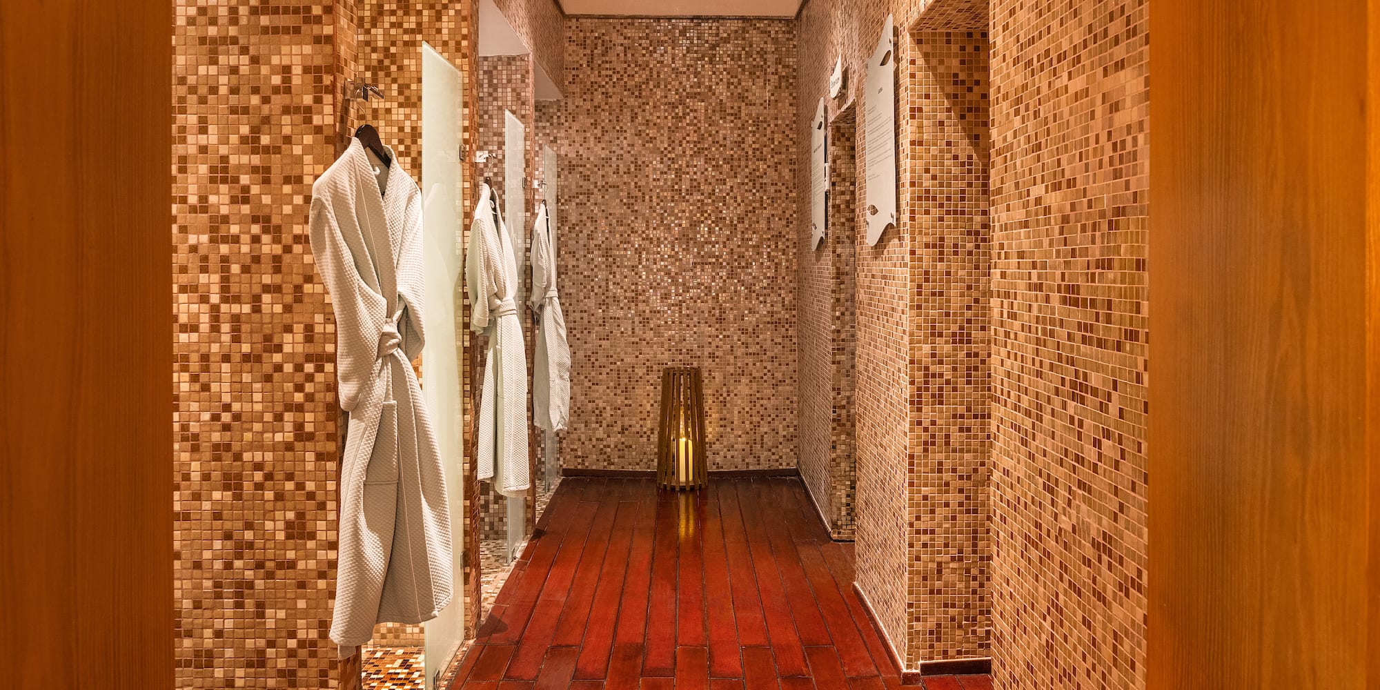 Luxury spa changing room with white bathrobes, mosaic walls, and polished wood floor.