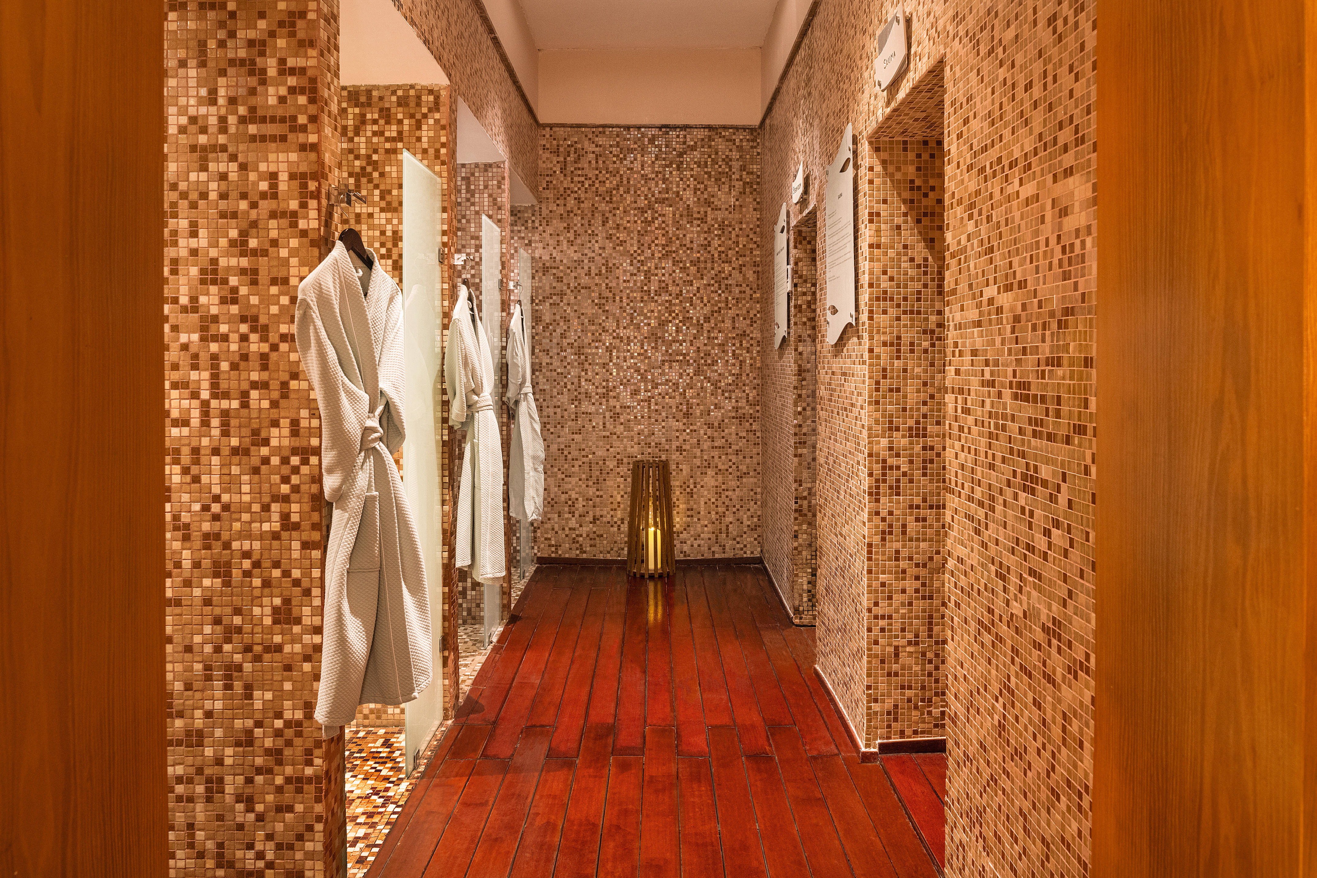 Luxury spa changing room with white bathrobes, mosaic walls, and polished wood floor.