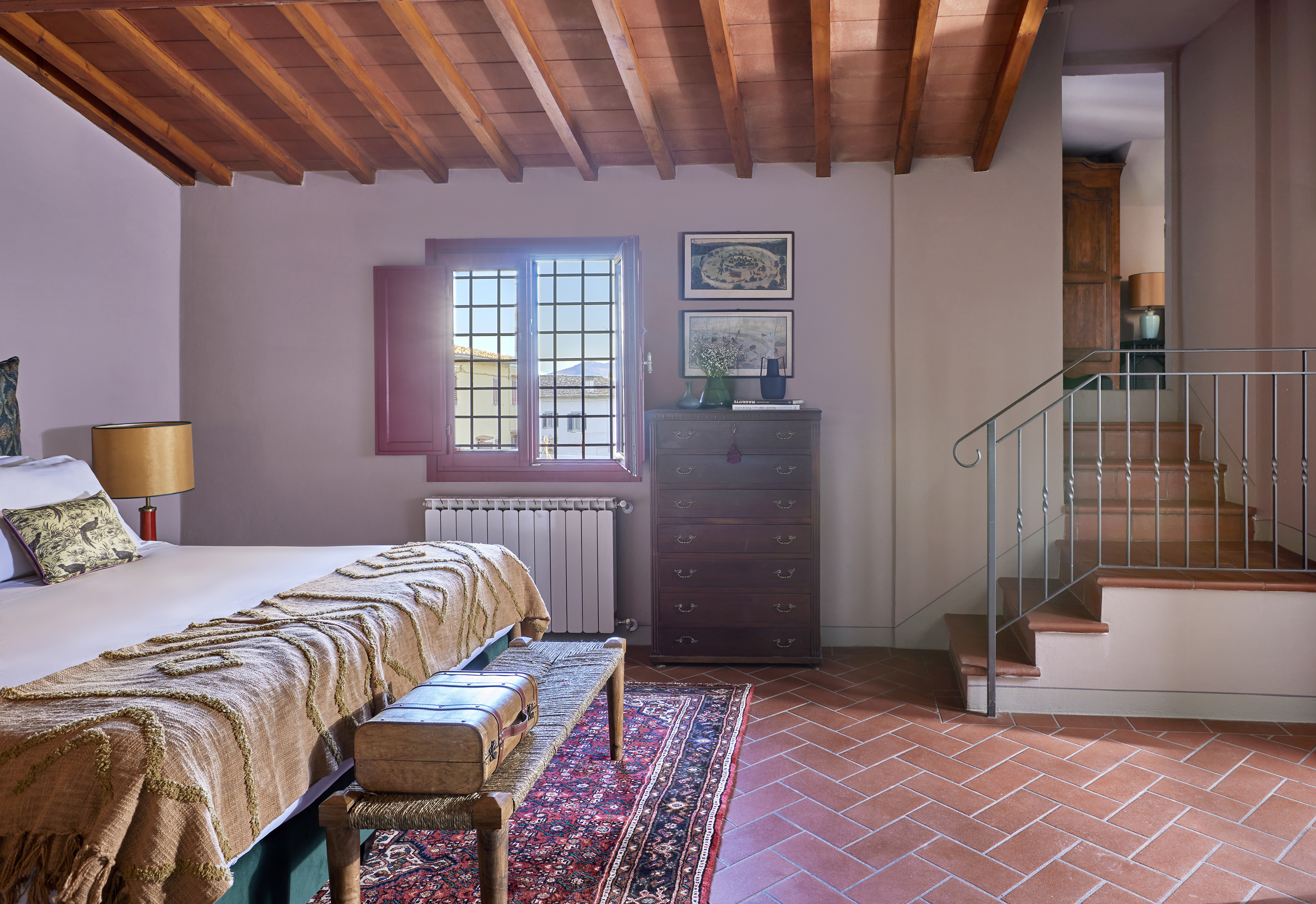 Terracotta floor bedroom with exposed beams, large bed, window, and stairs.
