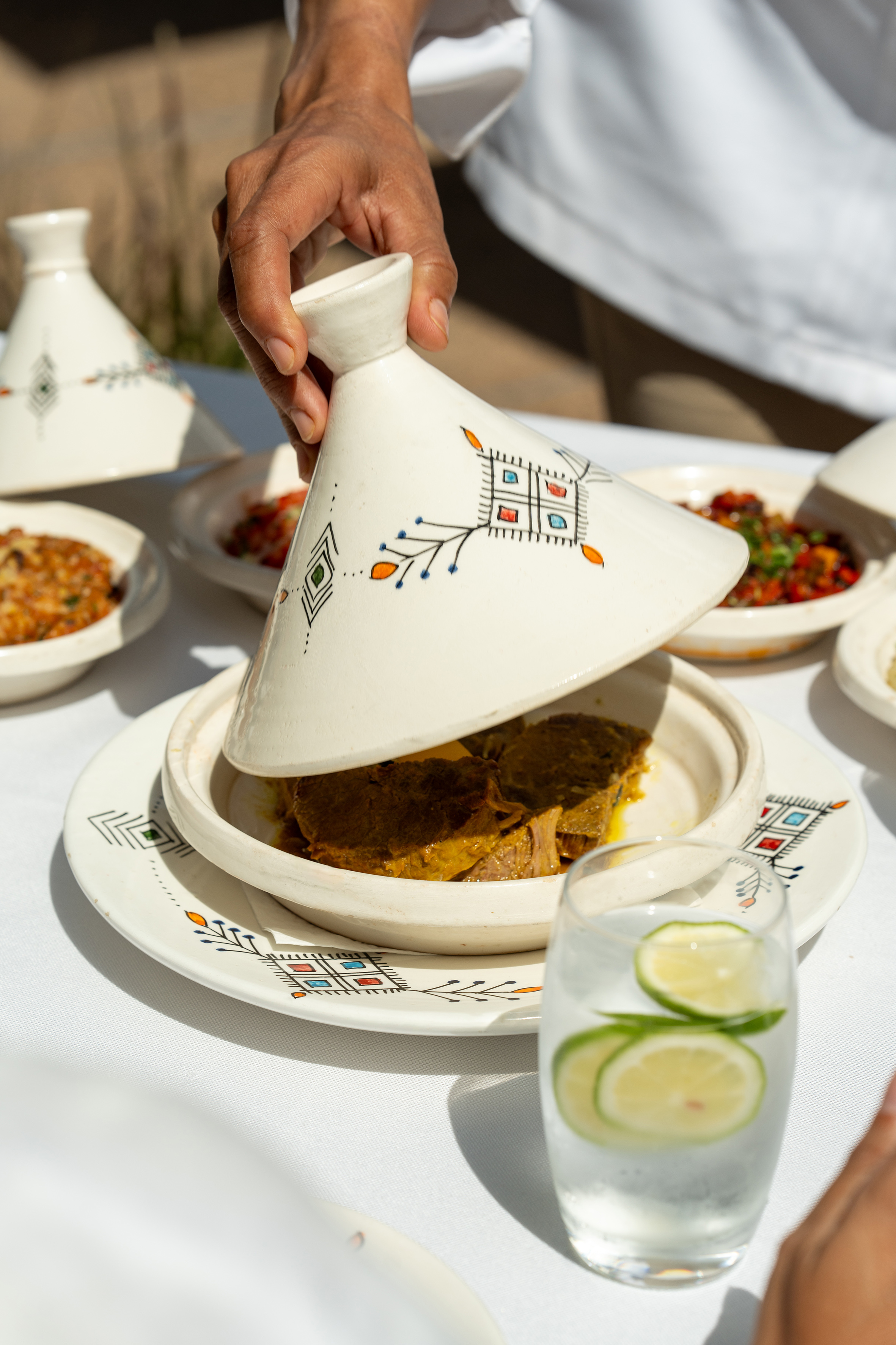 Hand lifting lid from a Moroccan tagine revealing a savory meat stew dish.