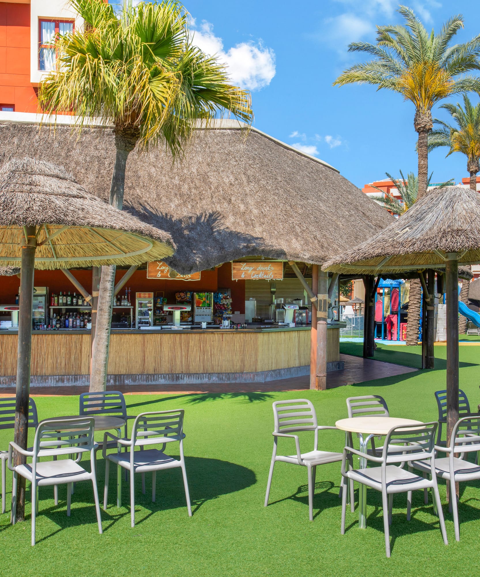 Resort thatched bar, outdoor seating on grass, palm trees, and hotel.