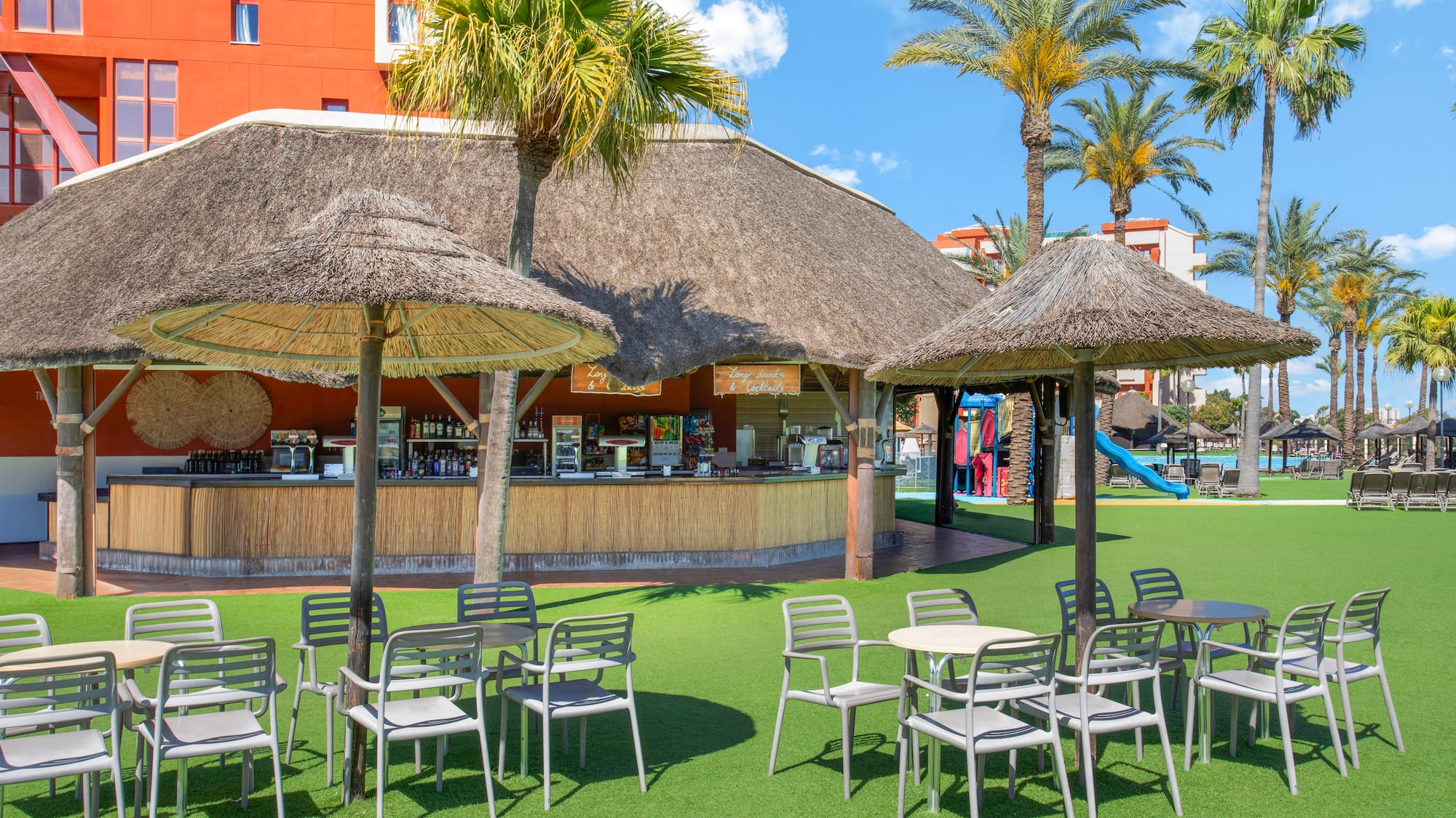 Tropical poolside bar with thatched roof, outdoor seating, and palm trees.