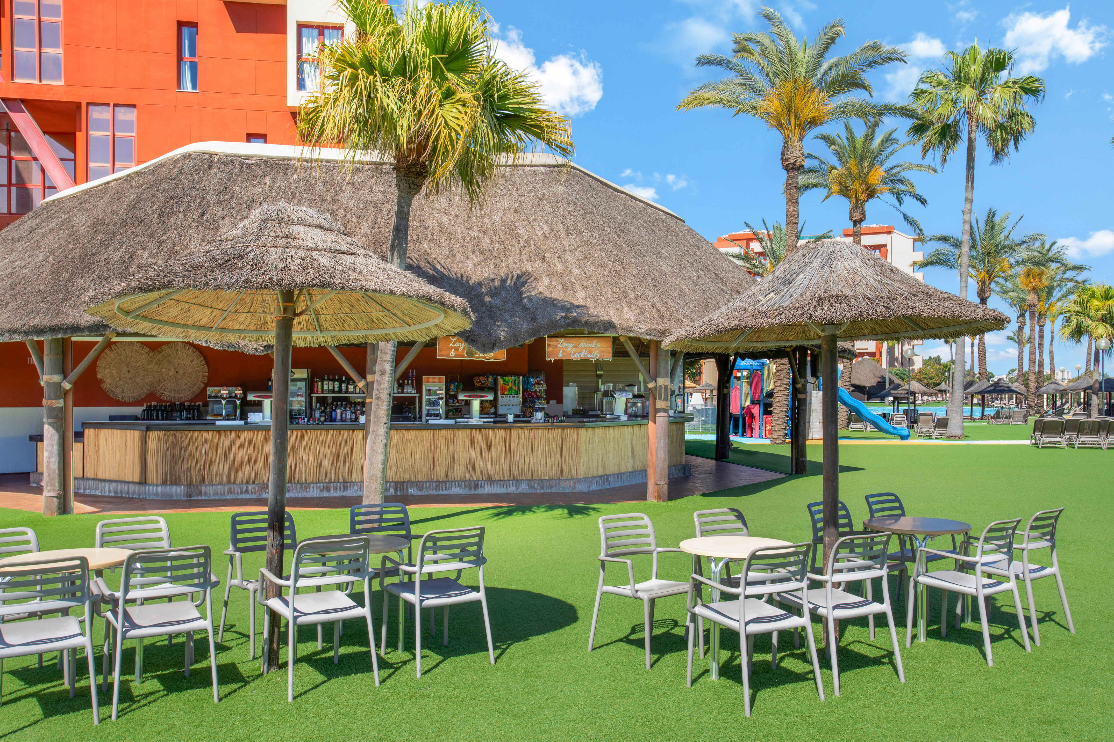 Resort thatched bar, outdoor seating on grass, palm trees, and hotel.
