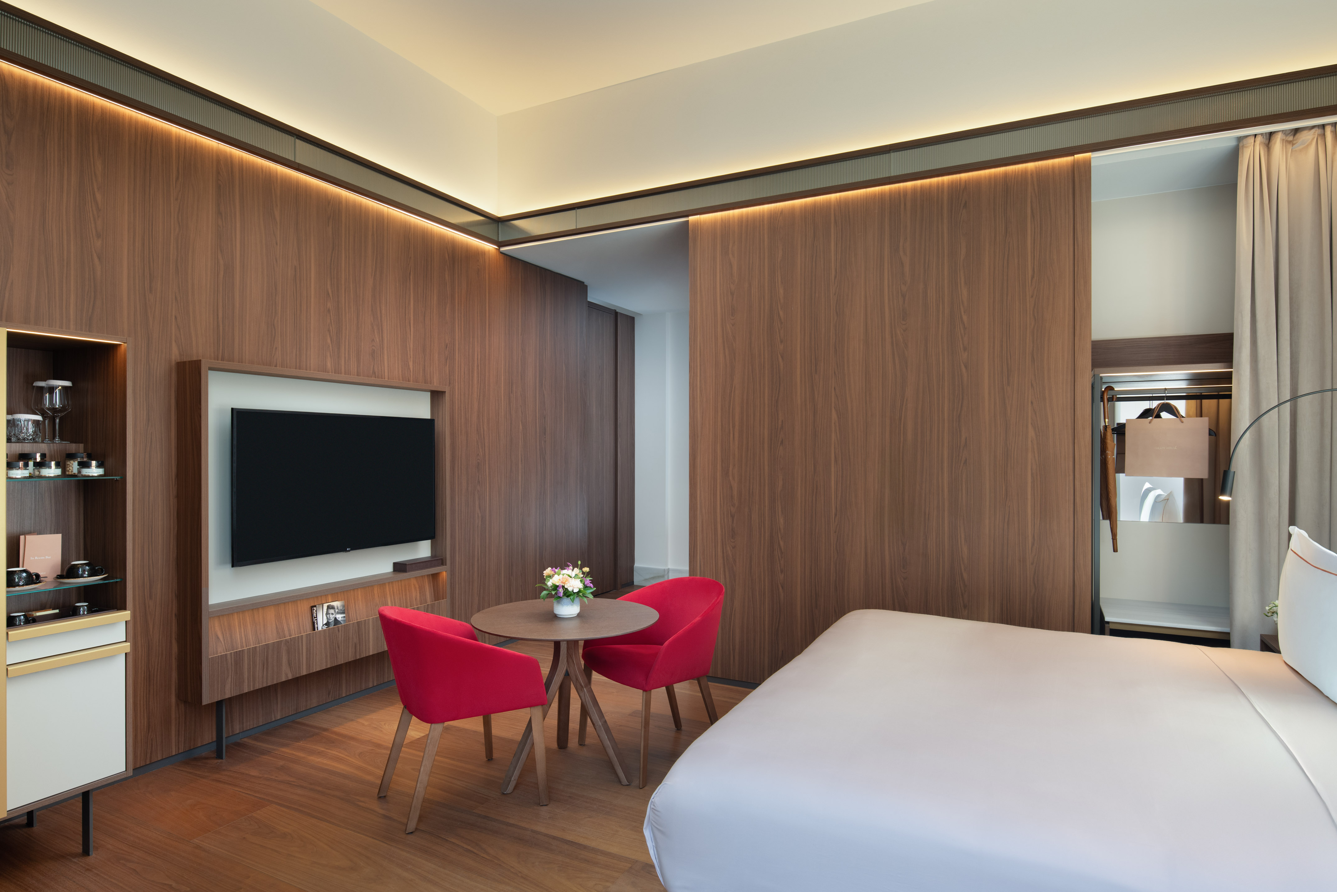 Contemporary hotel bedroom with wood walls, TV, red chairs, and white bed.