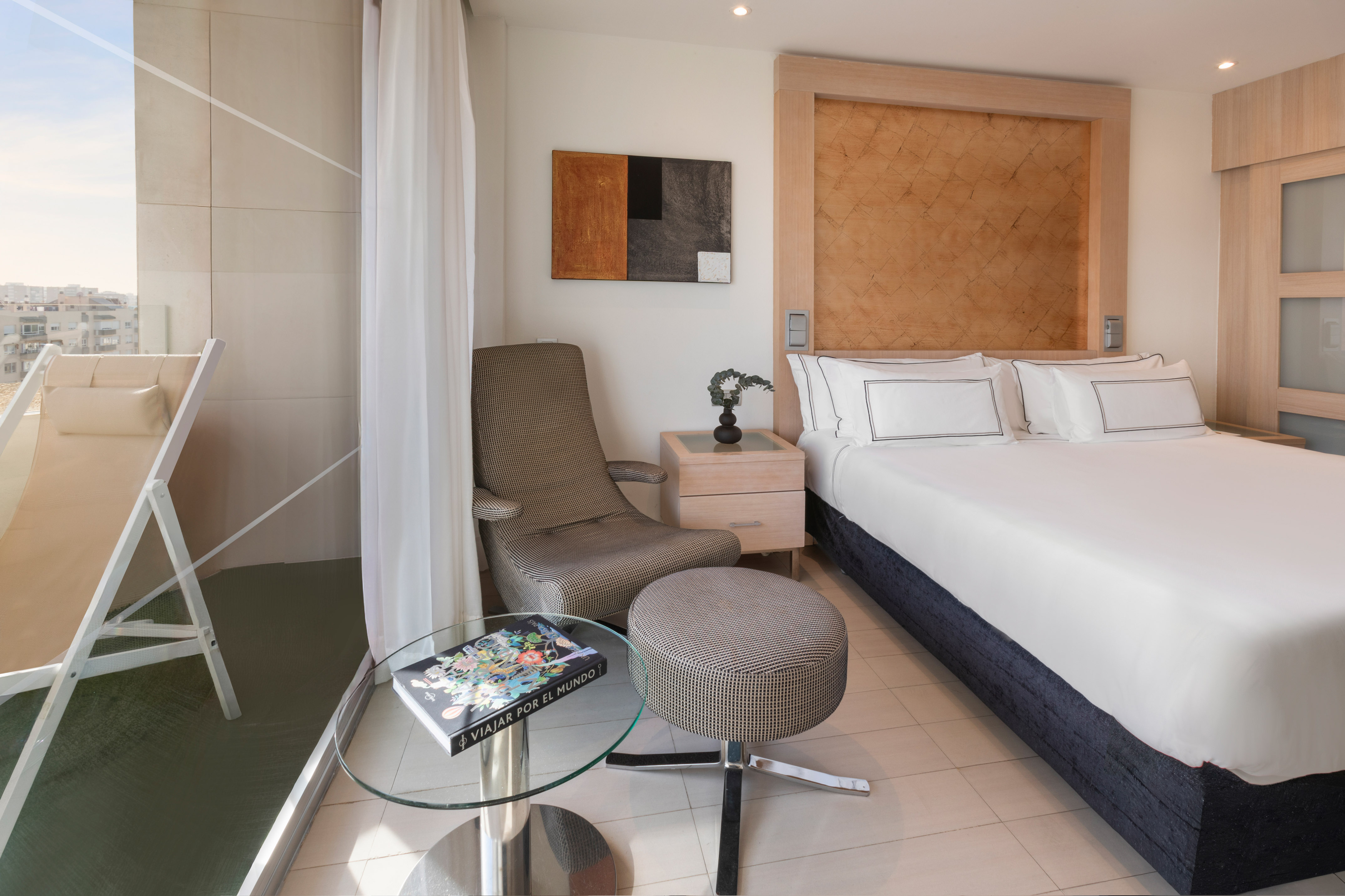 Modern hotel bedroom with bed, patterned armchair, and balcony cityscape.