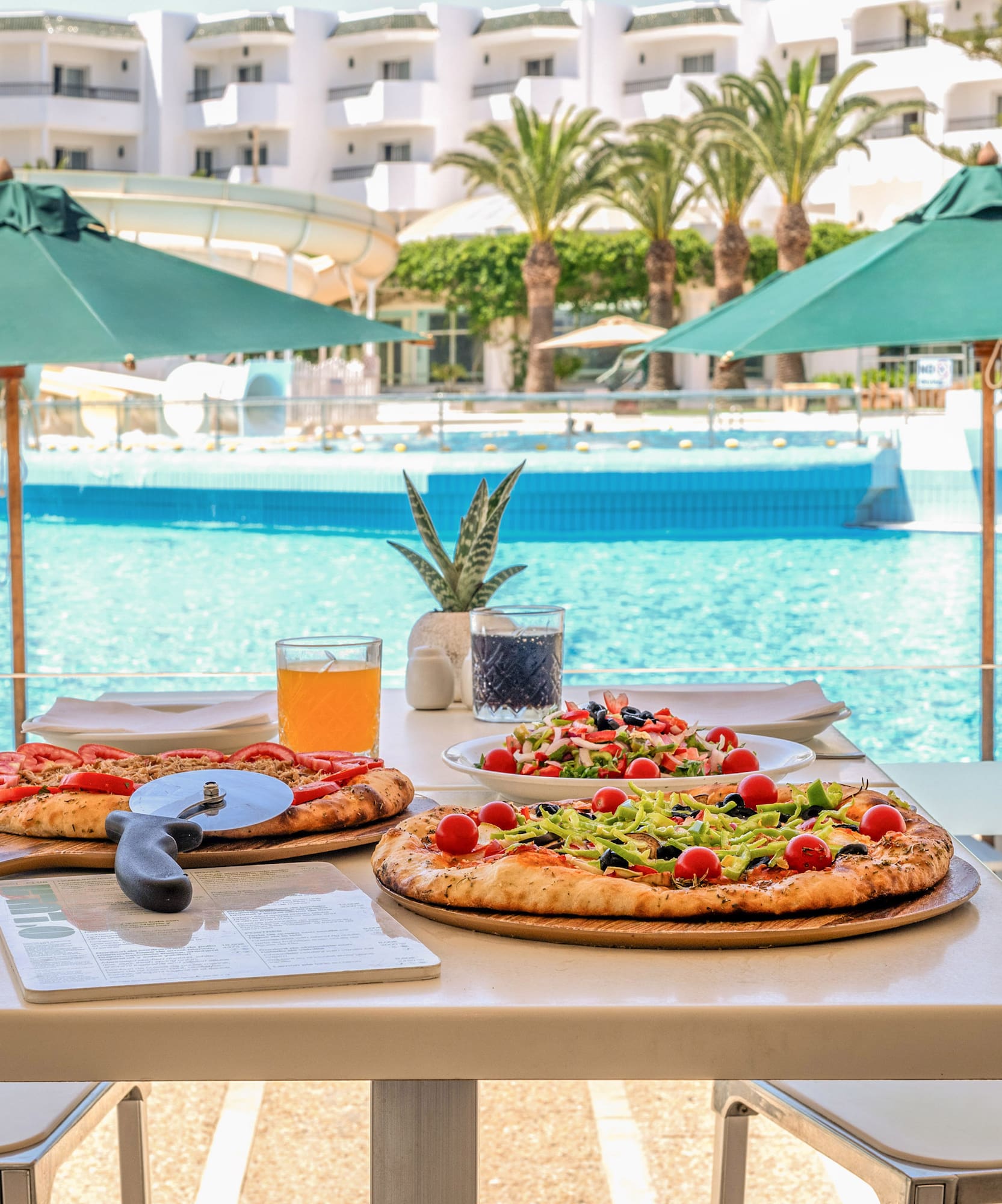 Pizzas y ensalada en mesa junto a piscina de hotel con toboganes.