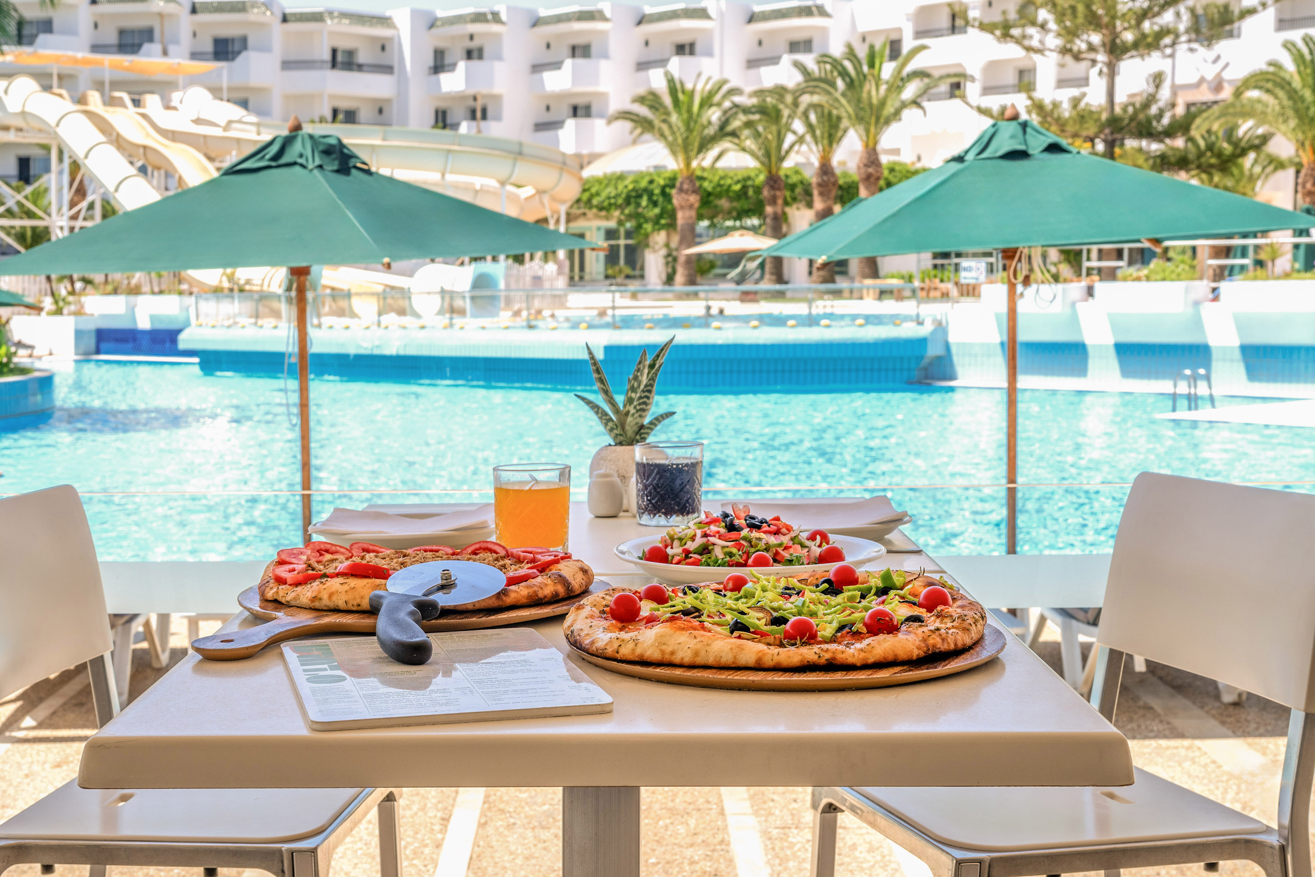 Pizzas y ensalada en mesa junto a piscina de hotel con toboganes.