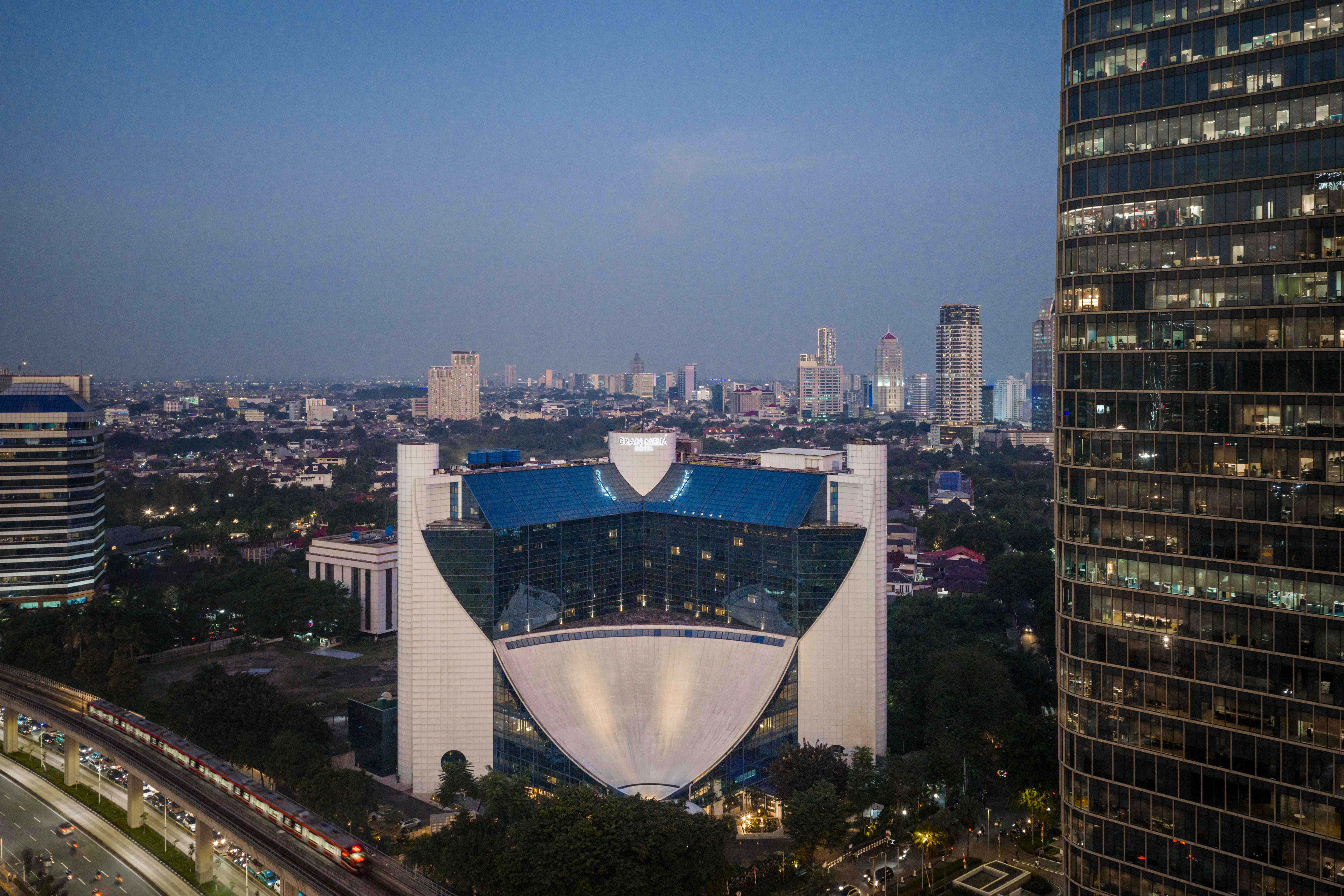 Gran Meliá Jakarta hotel, city skyline at dusk with train and skyscrapers.