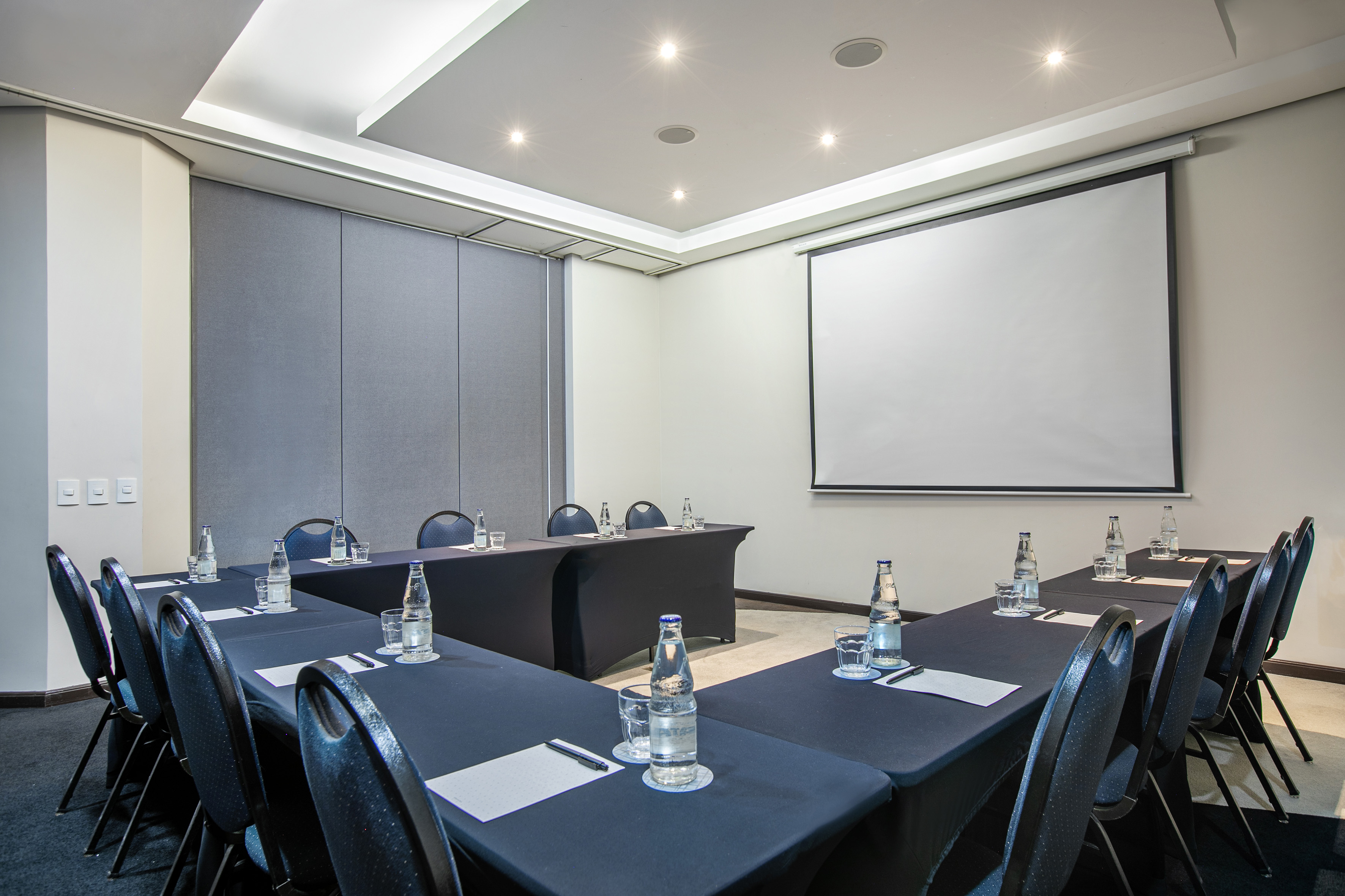Modern conference room with U-shaped tables, projector screen, water bottles.