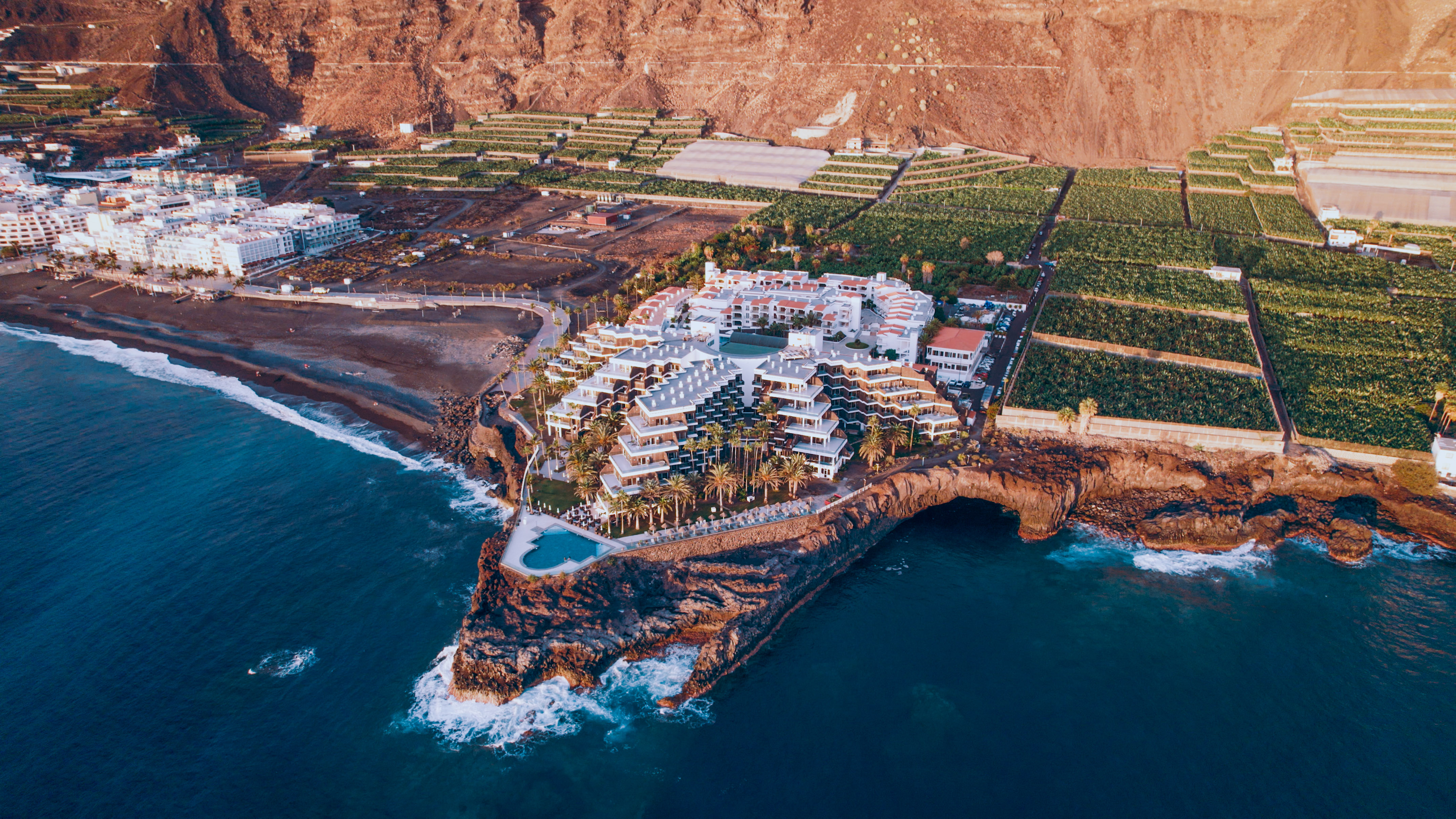 Coastal hotel on cliffs, black sand beach, ocean, and banana plantations.