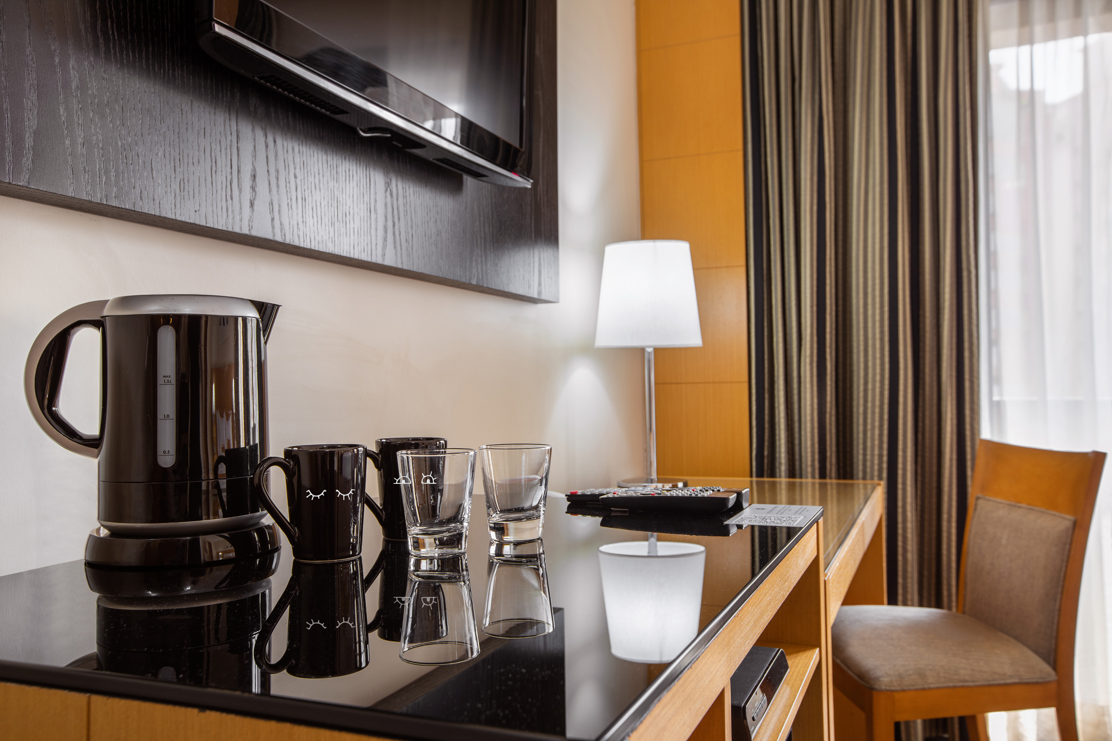 Hotel room desk area with electric kettle, coffee mugs, glasses, lamp, and TV.