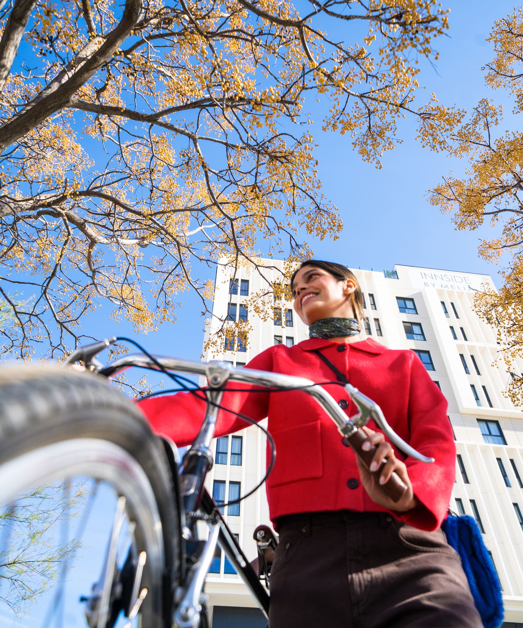 Smiling woman in red jacket with bicycle, autumn trees, and city building.
