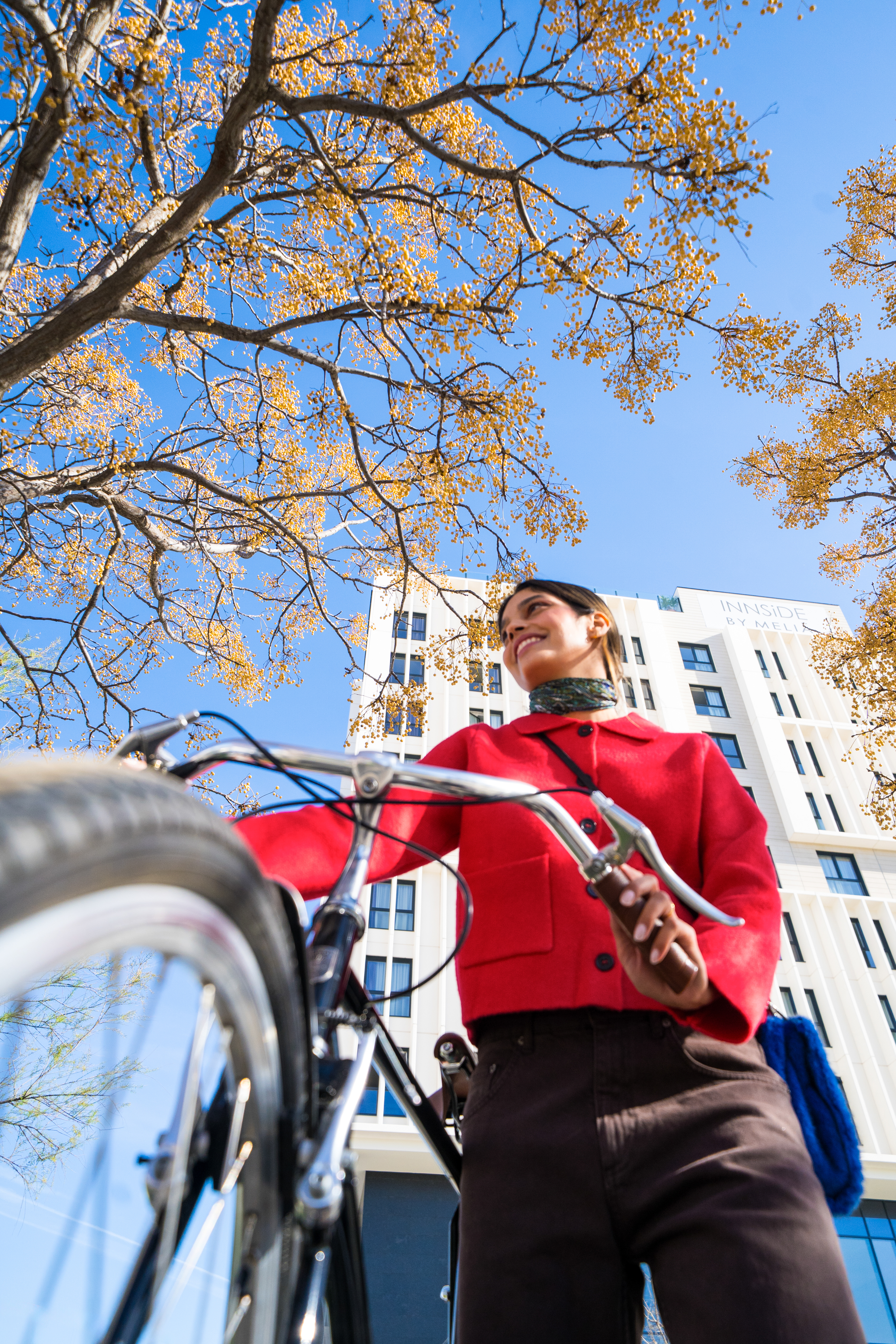 Smiling woman in red jacket with bicycle, autumn trees, and city building.