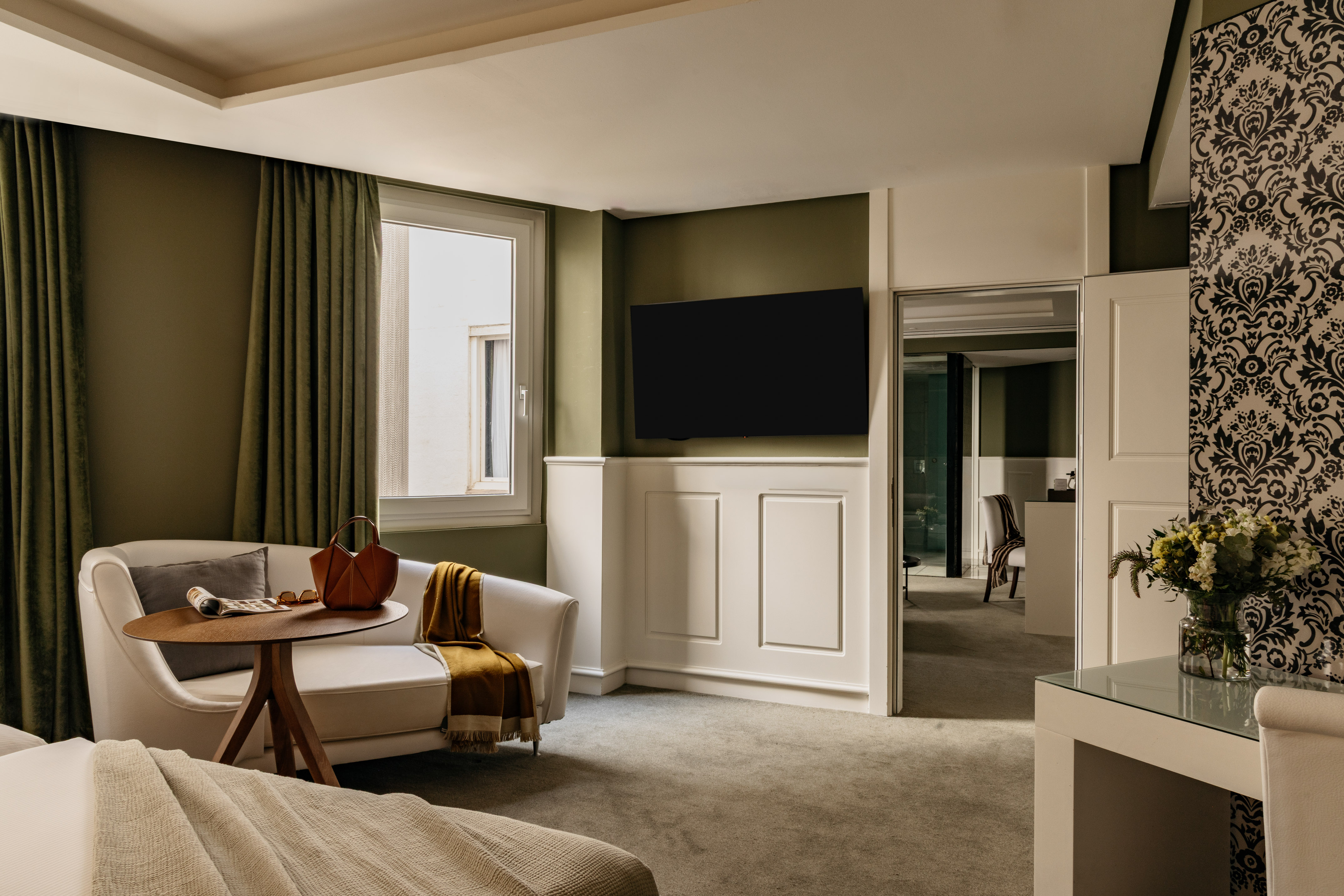 Suite de hotel con sofá blanco, TV, pared verde oliva y papel tapiz ornamental.