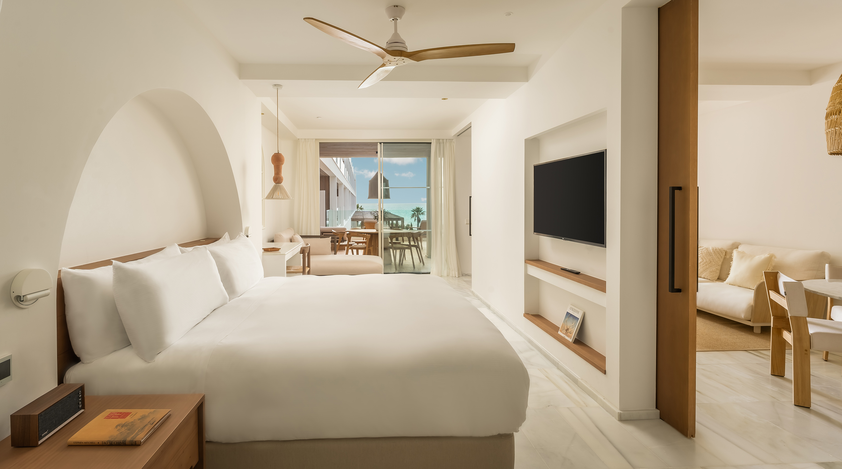 White hotel bedroom with king bed, ocean view balcony, and ceiling fan.