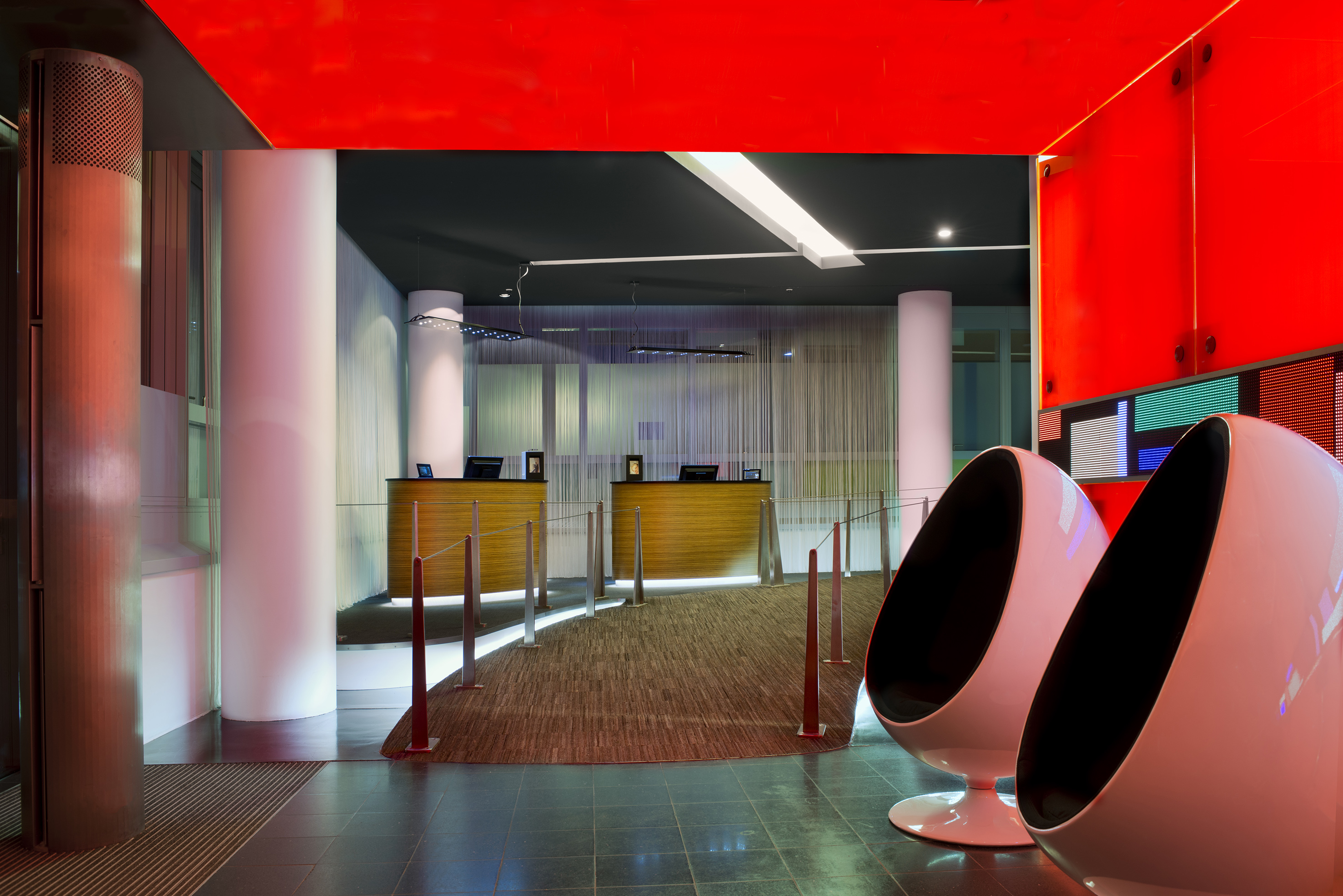 Modern hotel lobby with red ceiling, two reception desks, and white egg chairs.