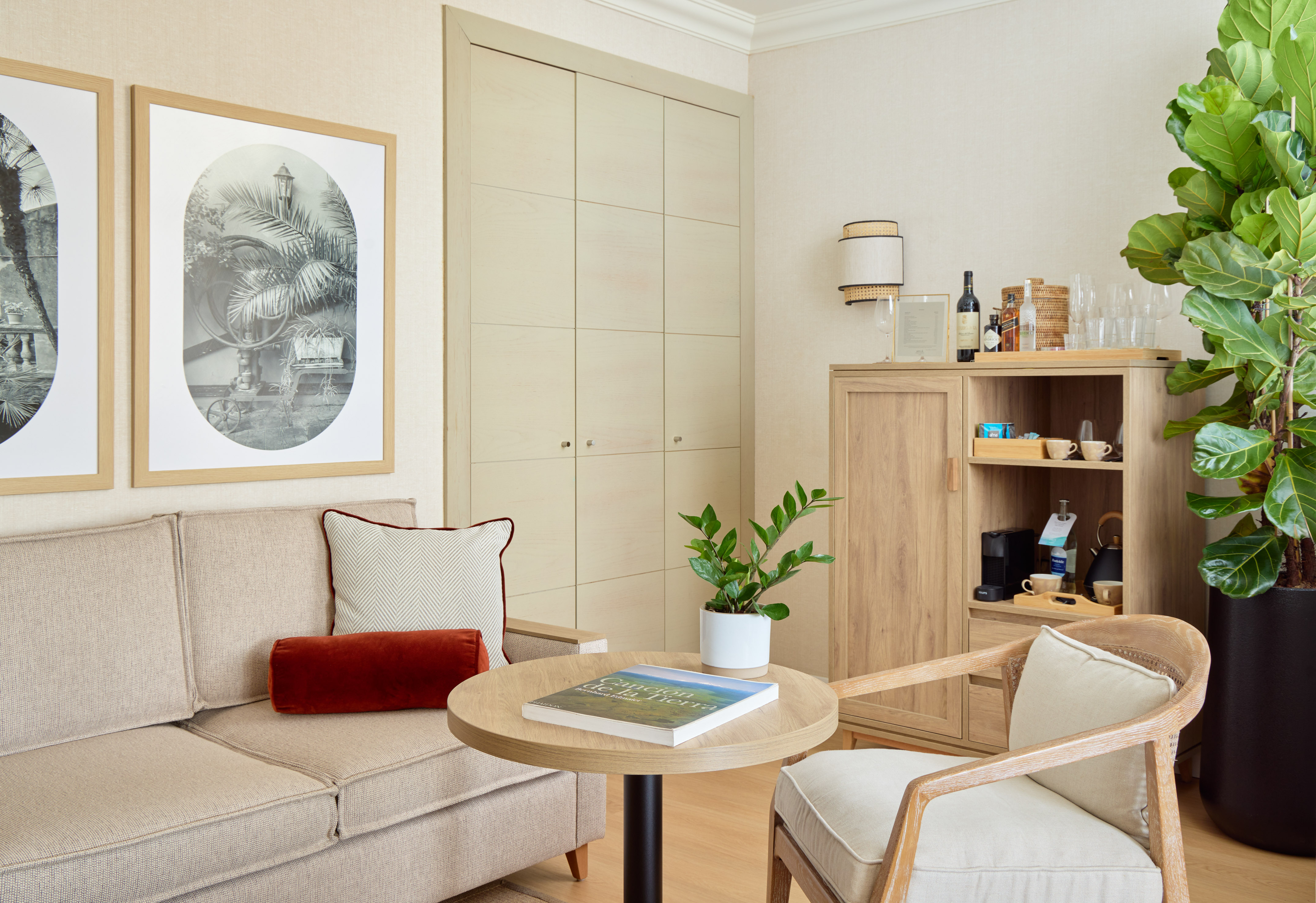 Hotel living area with beige sofa, wooden mini-bar, framed art, and large houseplant.