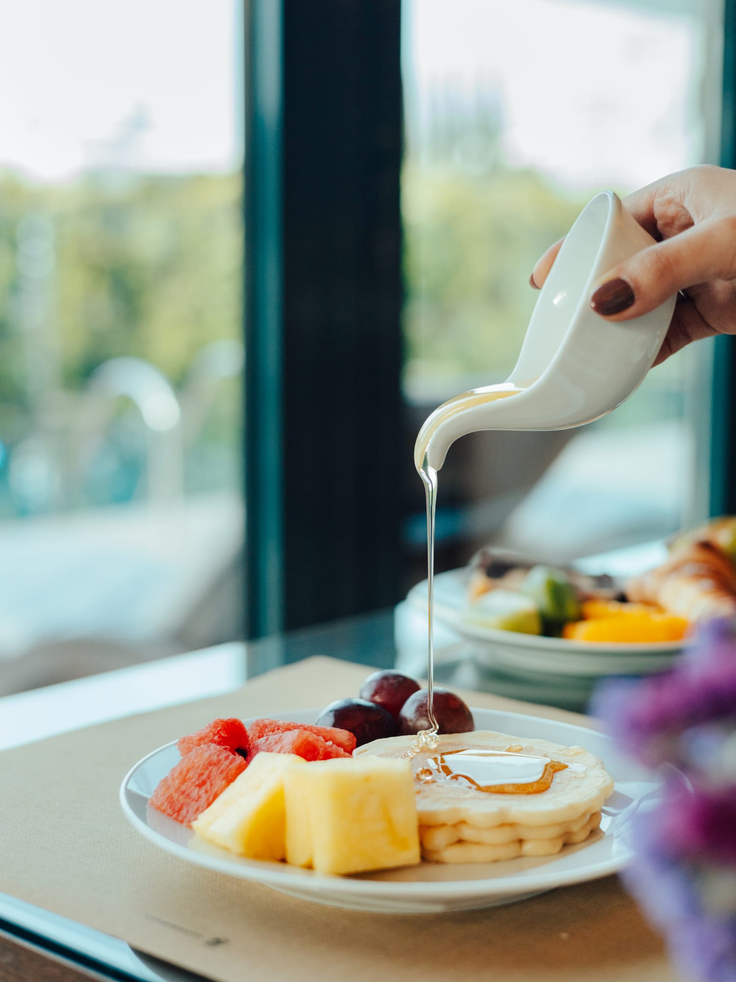Hand pouring golden syrup on pancakes with watermelon, pineapple, and grapes.