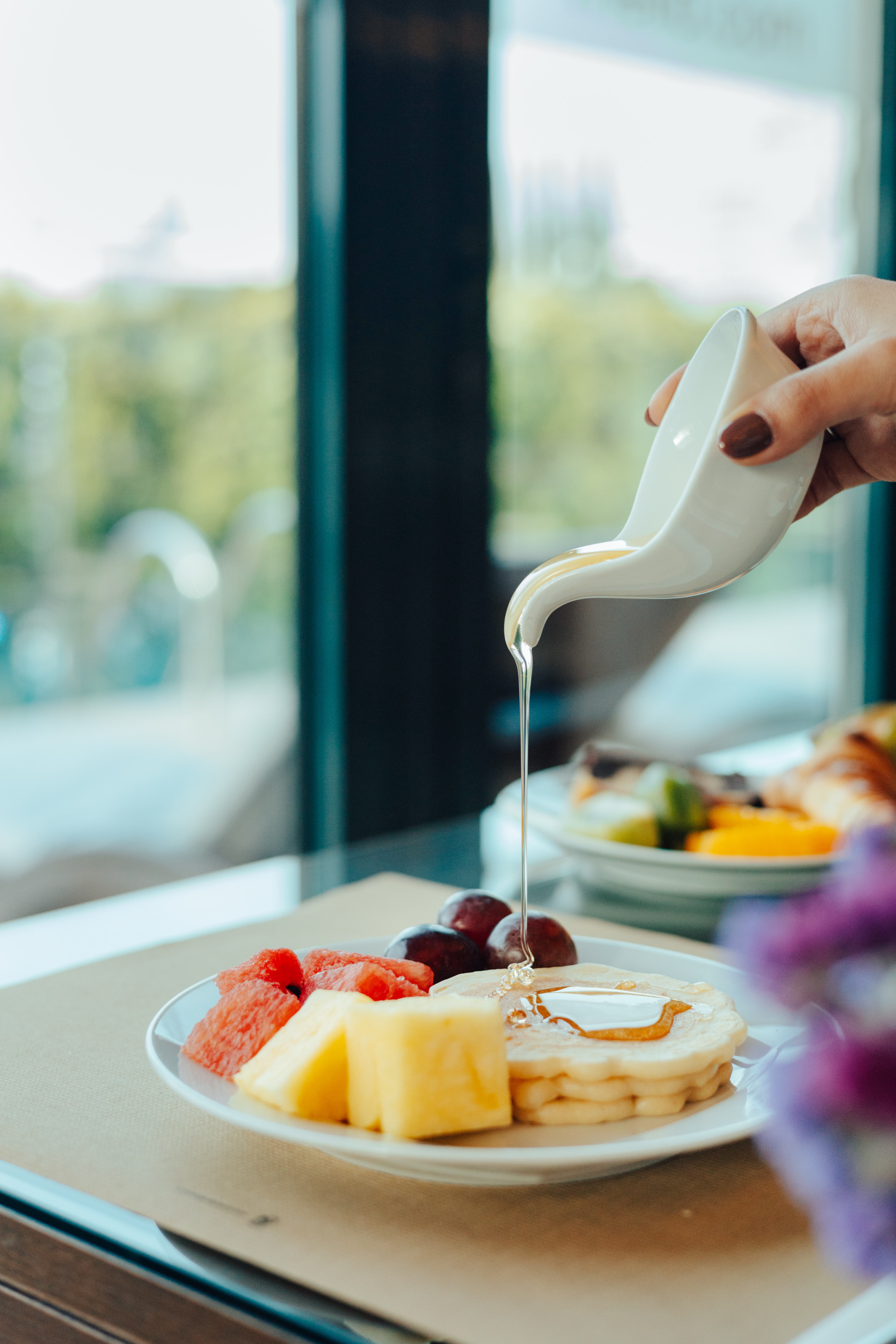 Hand pouring golden syrup on pancakes with watermelon, pineapple, and grapes.