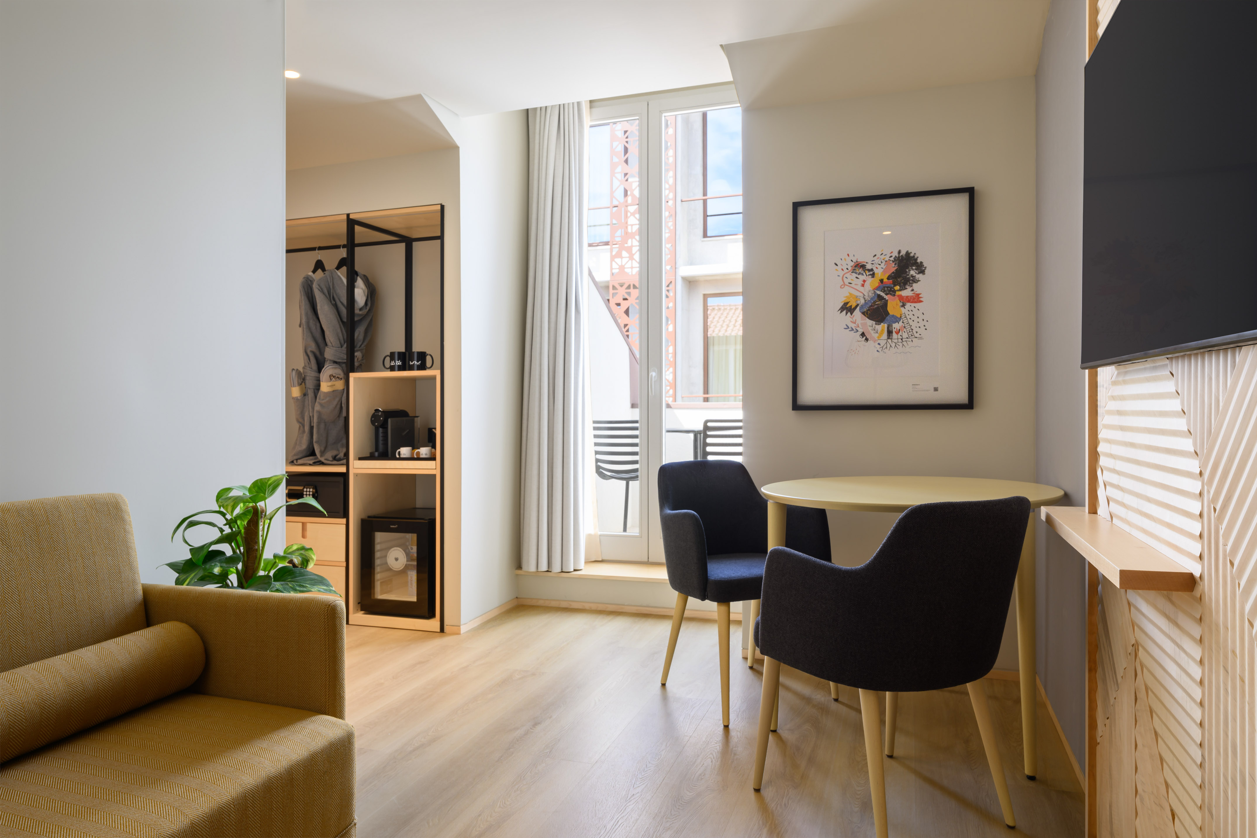Modern hotel room with dining set, sofa, open wardrobe, and balcony view.