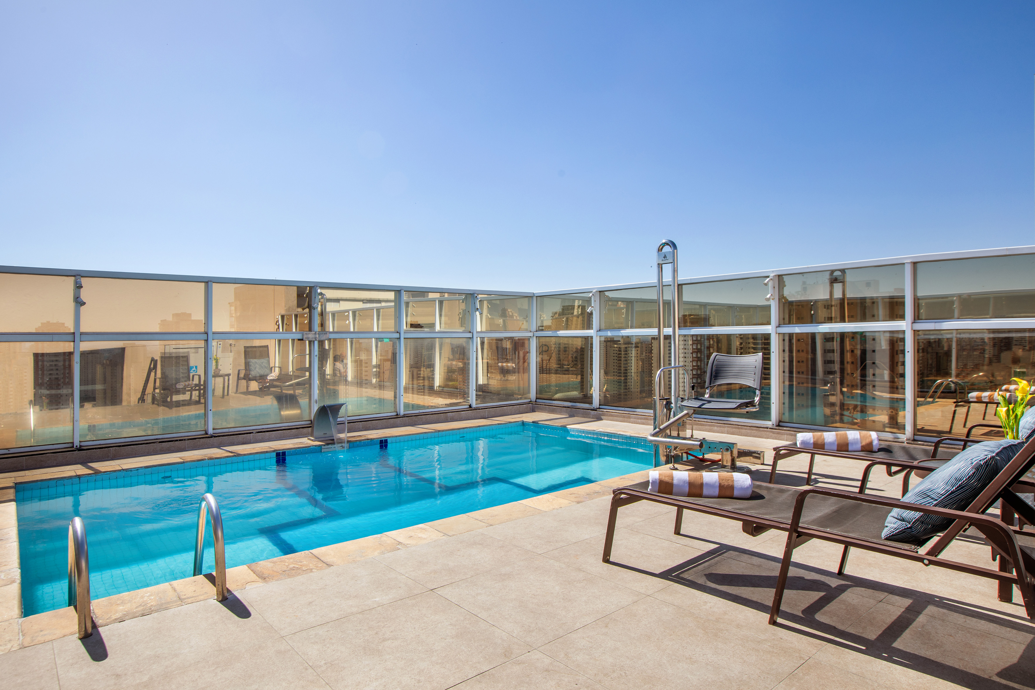Sunny rooftop pool with lounge chairs, city view, and clear blue water.