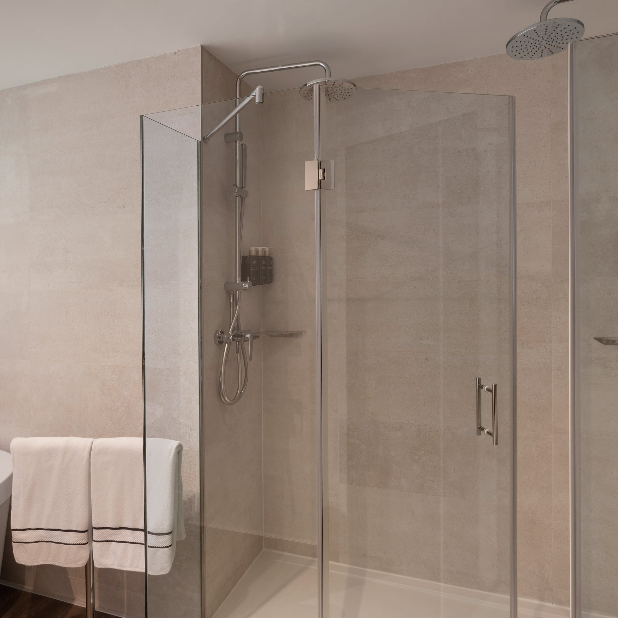 Baño moderno con ducha de cristal, bañera, azulejos claros y toallas.