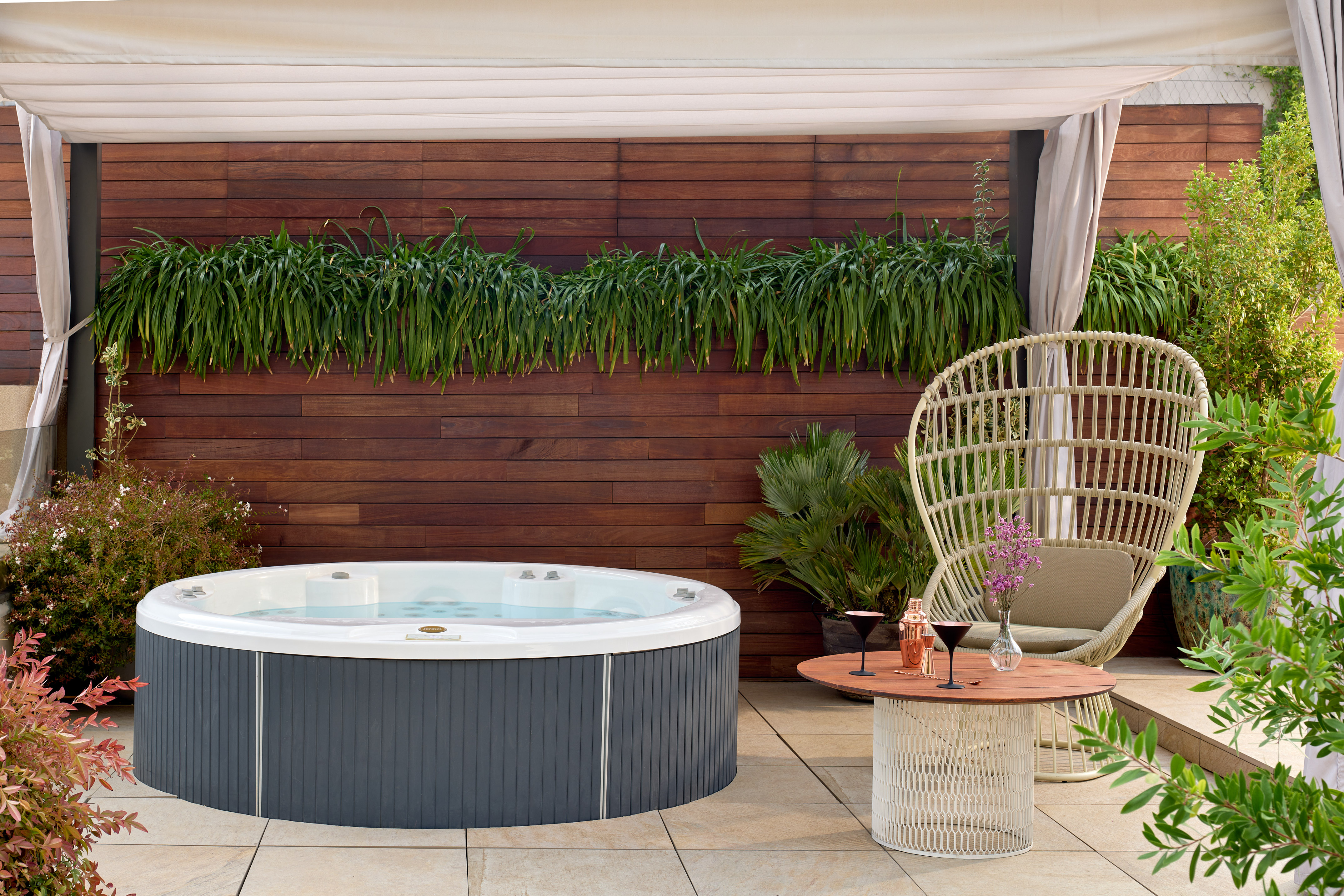 Outdoor hot tub on a modern patio with woven chair, cocktails, and lush greenery.